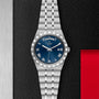 The TUDOR Royal watch features a stainless steel case, blue dial with Roman numerals, day and date display, and an integrated bracelet, displayed against a textured black and red background.