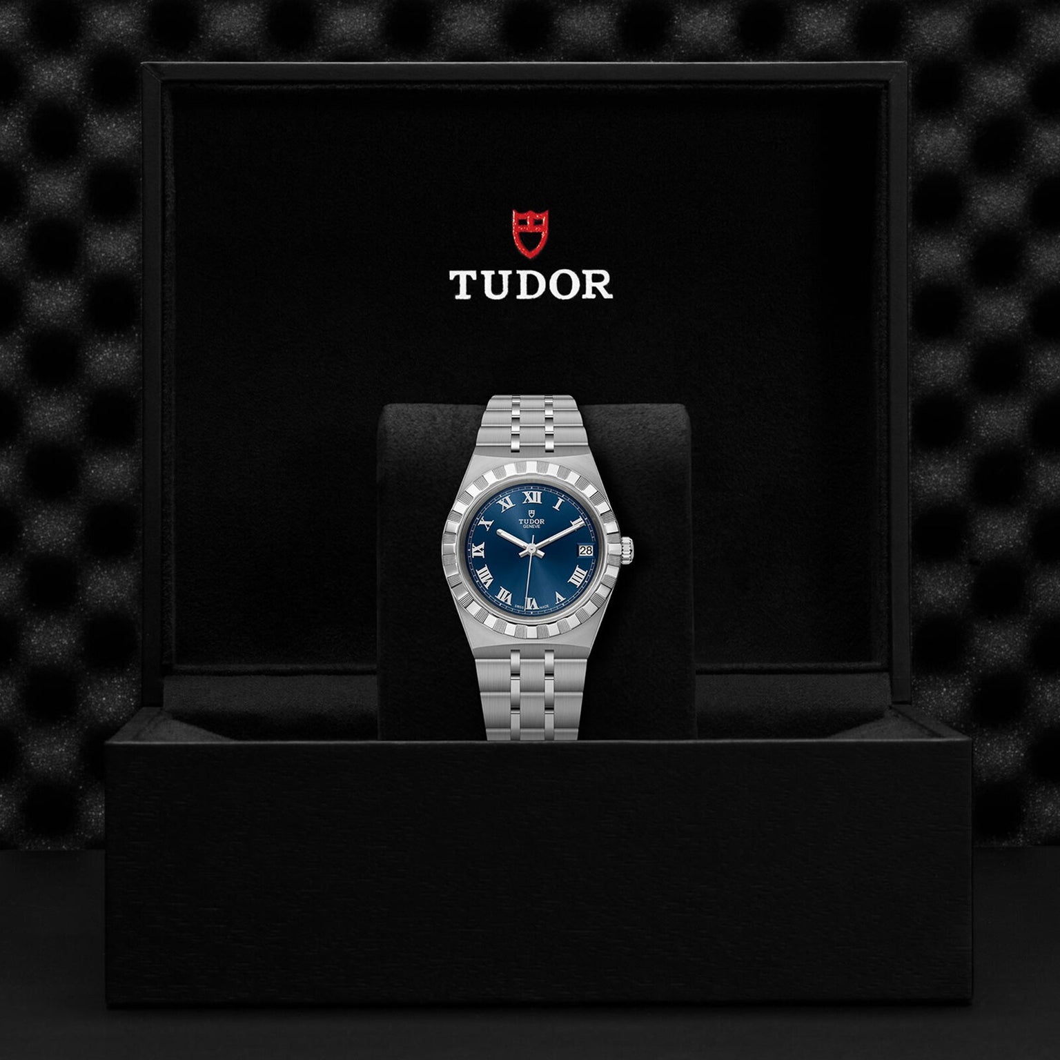 A TUDOR Royal watch with a blue dial and silver metal bracelet stands upright in a black Tudor-branded presentation box.