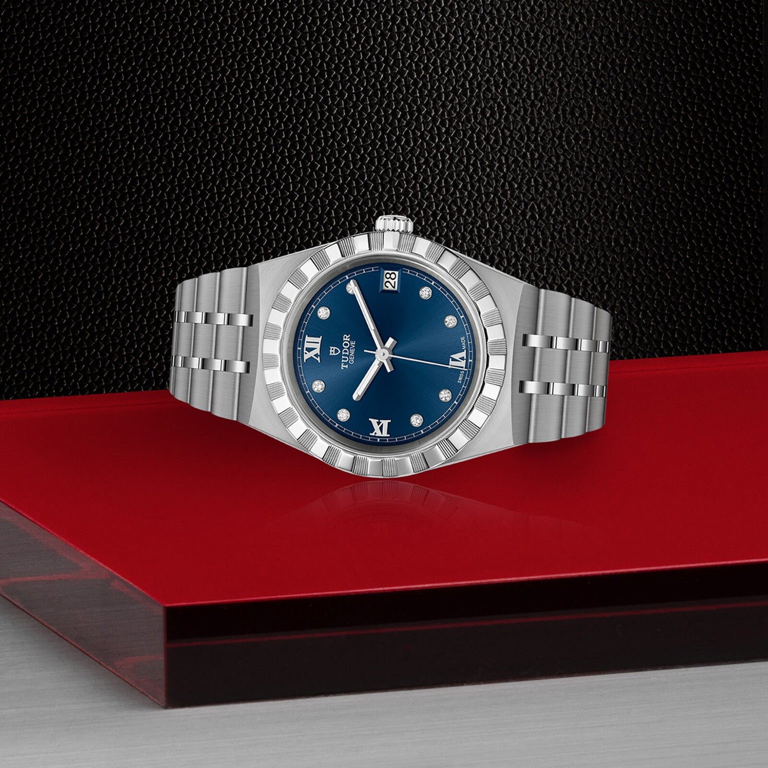 A TUDOR Royal automatic sport-chic watch in silver with a blue dial, diamond markers, and metal bracelet is showcased on a red and gray platform against a textured black backdrop.