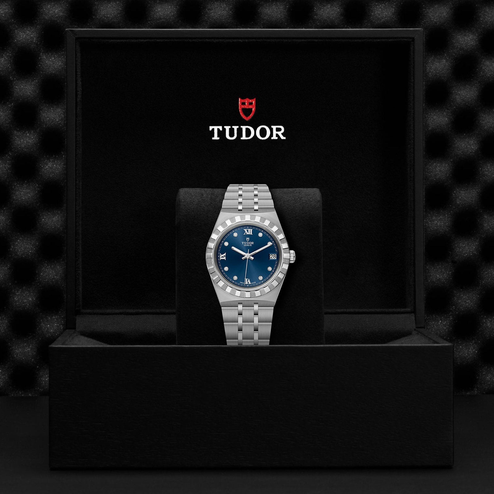 A TUDOR Royal wristwatch with a blue dial is showcased on a stand inside an open black Tudor watch box, highlighting the refined elegance of this automatic sport-chic timepiece.