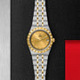 A TUDOR Royal wristwatch with a gold dial, Roman numerals, date display, and a stainless steel and gold bracelet is displayed on a textured black surface.