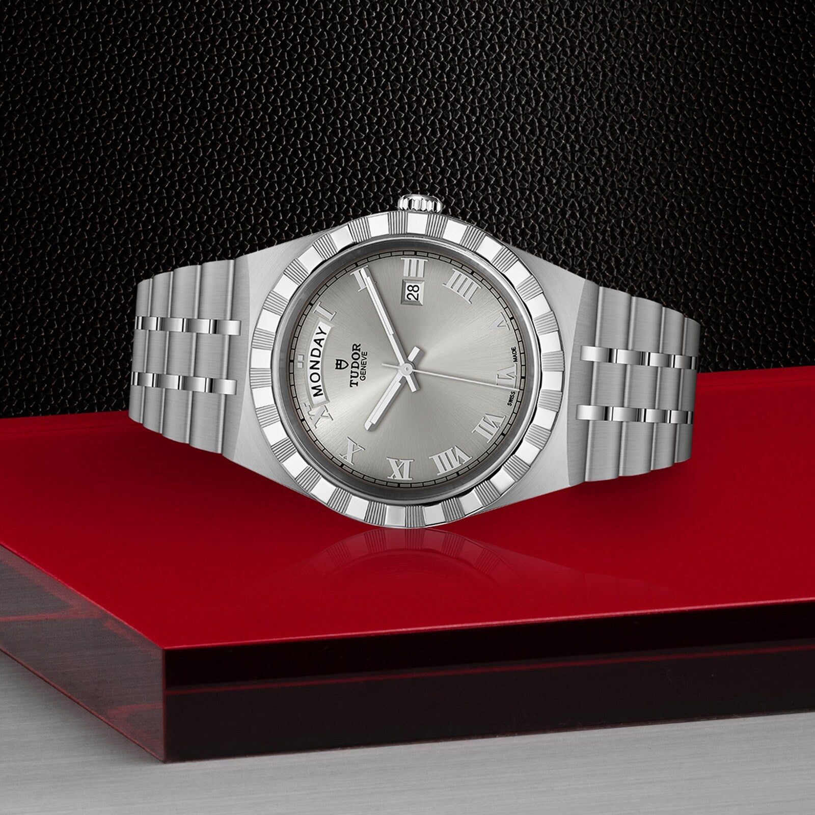 A TUDOR Royal wristwatch in silver, featuring an integrated bracelet, Roman numeral markers, and day-date displays, is displayed on a red and black geometric surface.