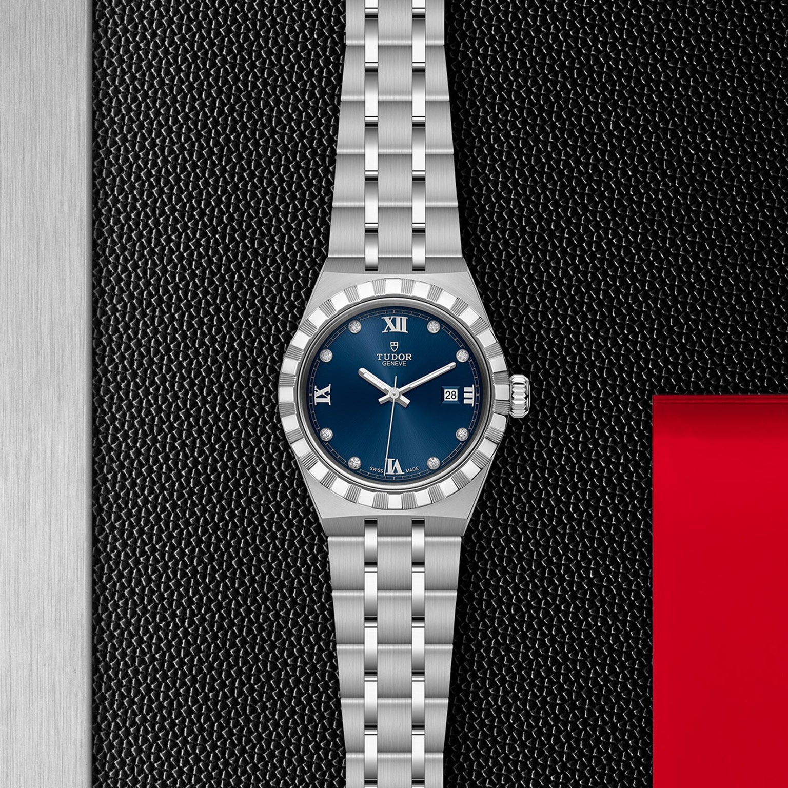 A TUDOR Royal wristwatch with a blue dial, Roman numerals, date display, and integrated bracelet rests on a textured black and metallic background with a red accent—showcasing the sport-chic spirit of this automatic watch.