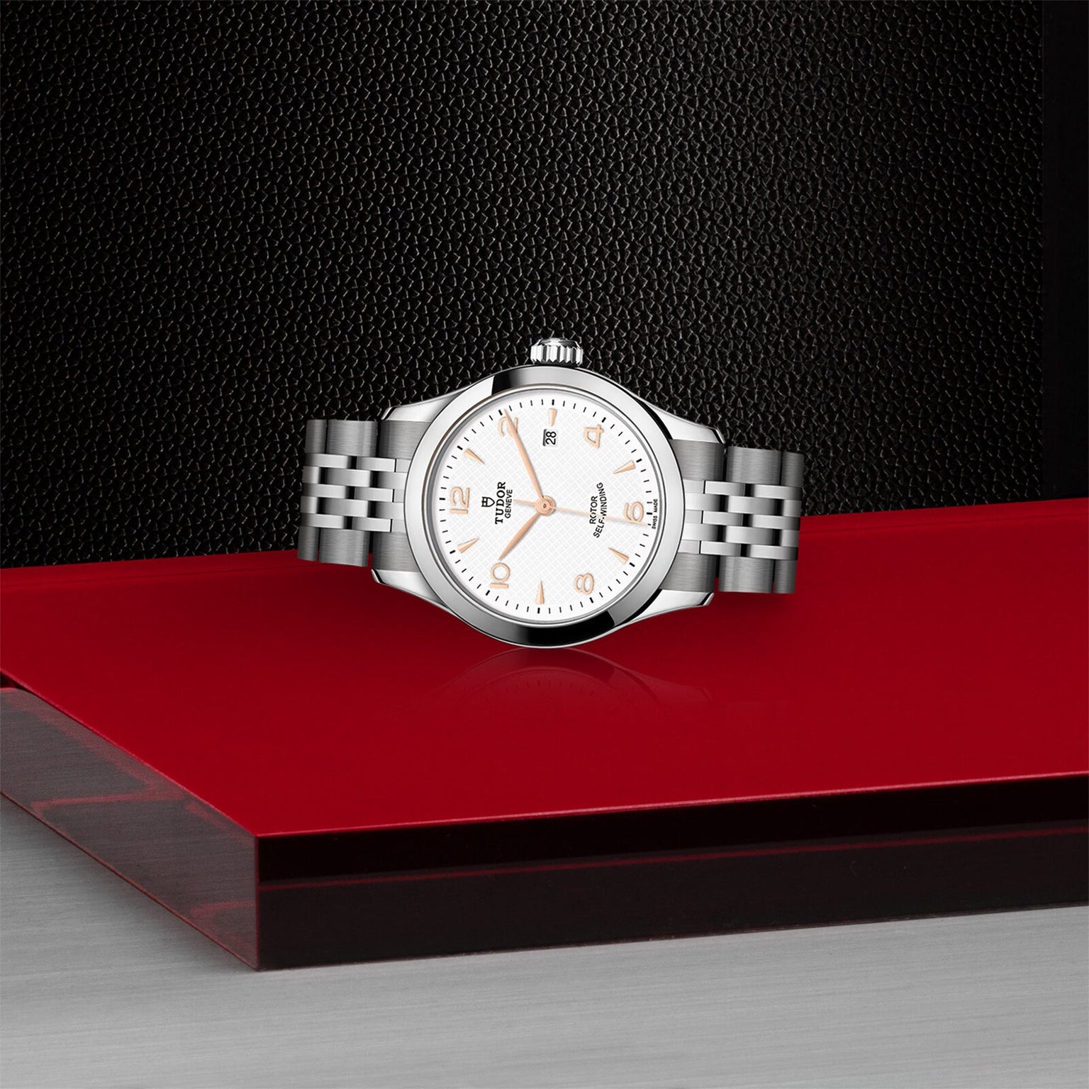 The TUDOR 1926, a silver wristwatch with a white dial and metal bracelet, is showcased on a glossy red and black platform against a textured black background.