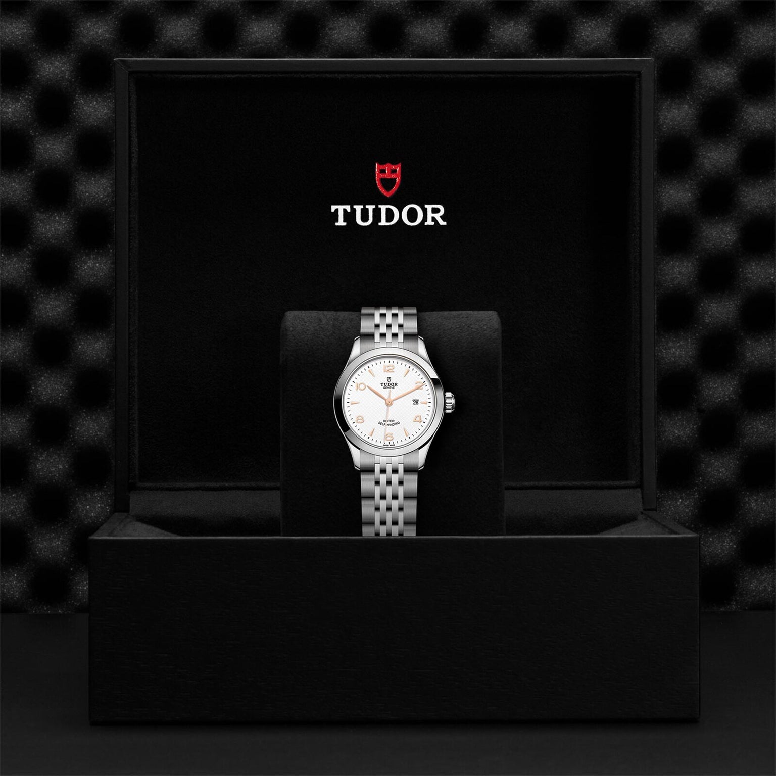 A TUDOR 1926 silver wristwatch with a white dial, known for its classic elegance, is shown in an open black presentation box featuring the TUDOR logo inside the lid.