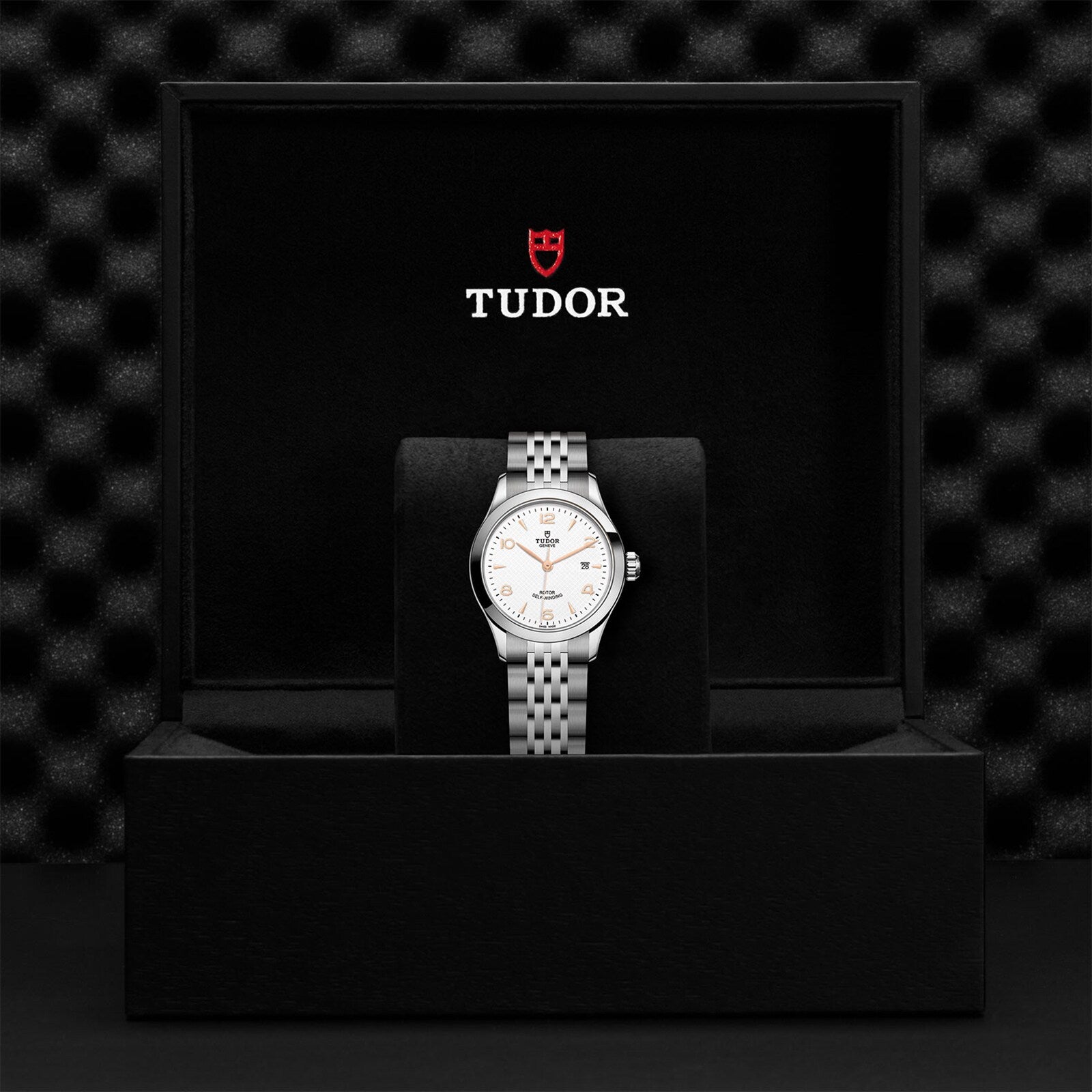 A TUDOR 1926 silver wristwatch with a white dial, known for its classic elegance, is shown in an open black presentation box featuring the TUDOR logo inside the lid.