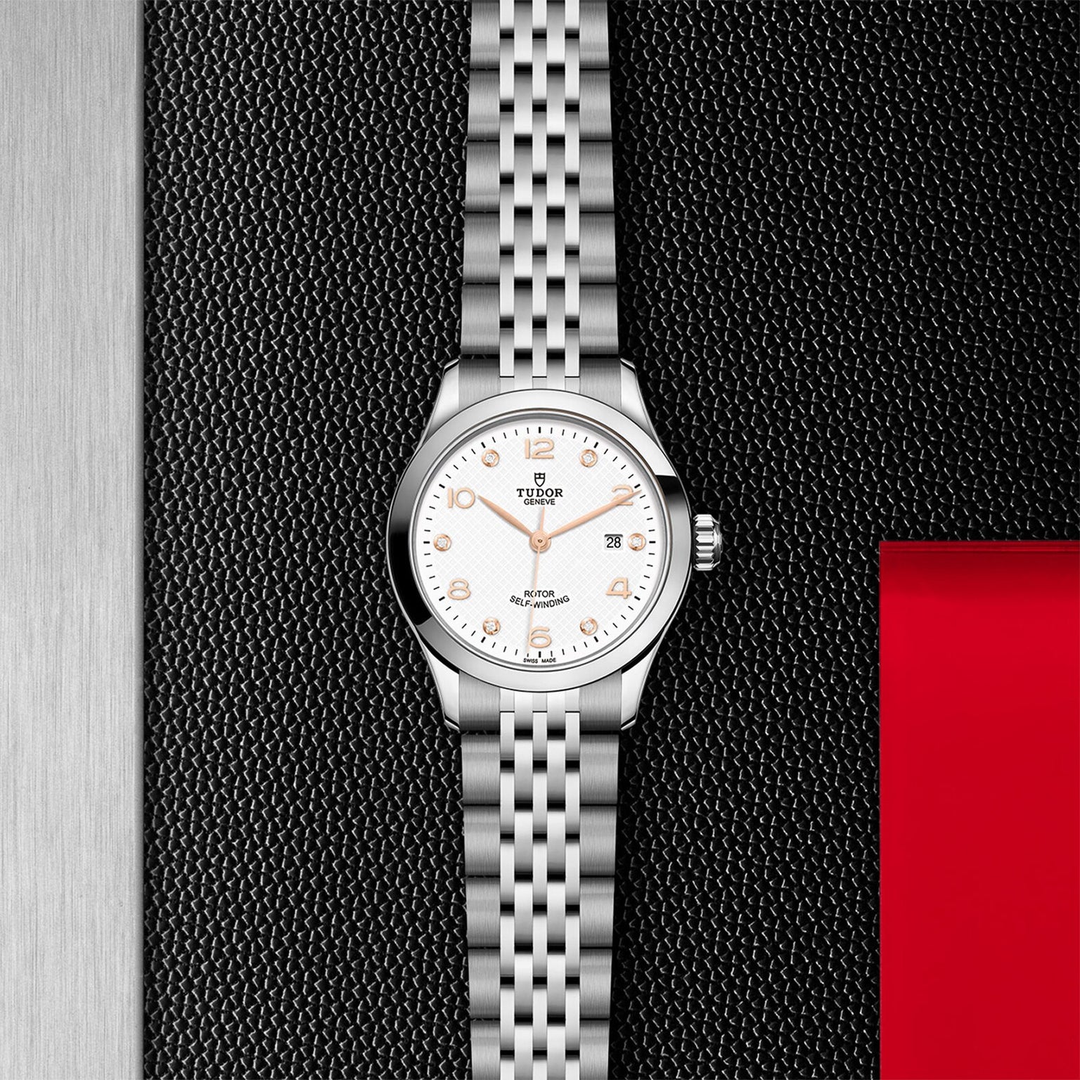 The TUDOR 1926 wristwatch features a silver case, white dial, gold hour markers, and a metal link bracelet, shown on a textured black surface beside red and metallic panels—a timeless choice for mechanical watch lovers.