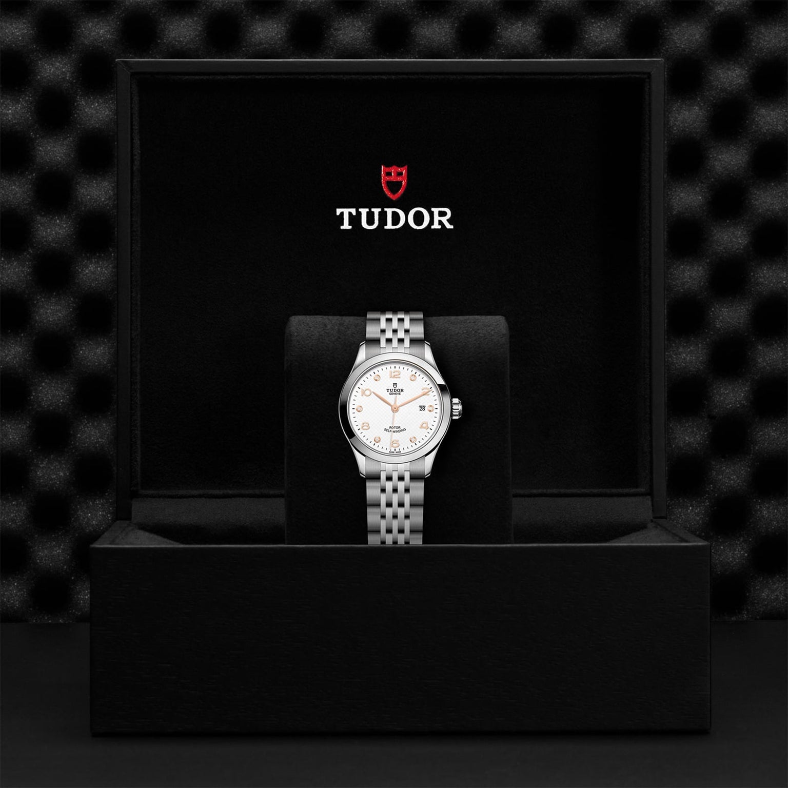 A TUDOR 1926 silver wristwatch with a white dial and metal bracelet is displayed upright in an open black box bearing the TUDOR logo.