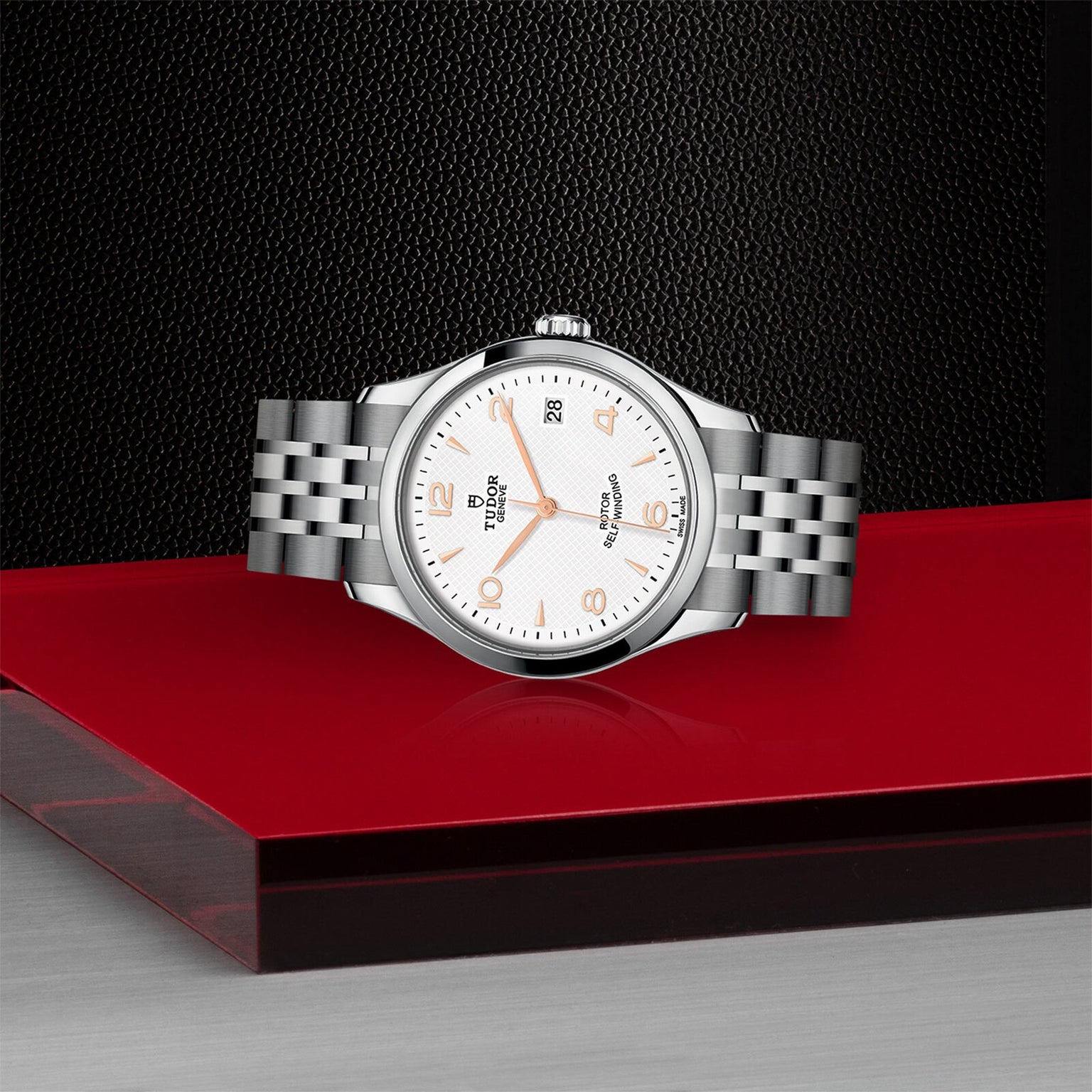 A TUDOR 1926 wristwatch with a silver metal bracelet and white dial is showcased on a glossy red and black surface, set against a textured black background.