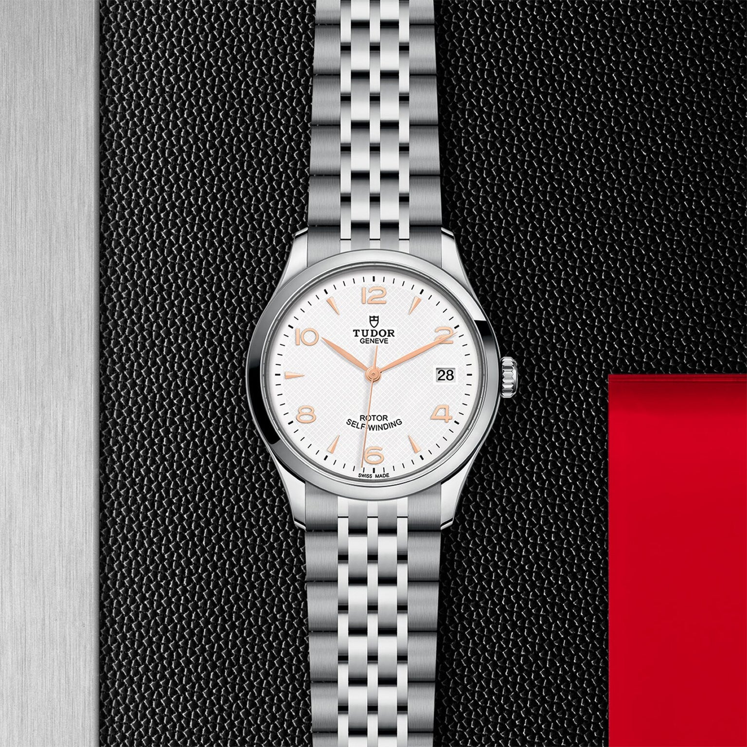 The TUDOR 1926 wristwatch features a classic silver design with a white dial, orange numerals, date window, and metal link bracelet, displayed on a black and silver textured background with a red accent—a timeless choice for mechanical watch enthusiasts.