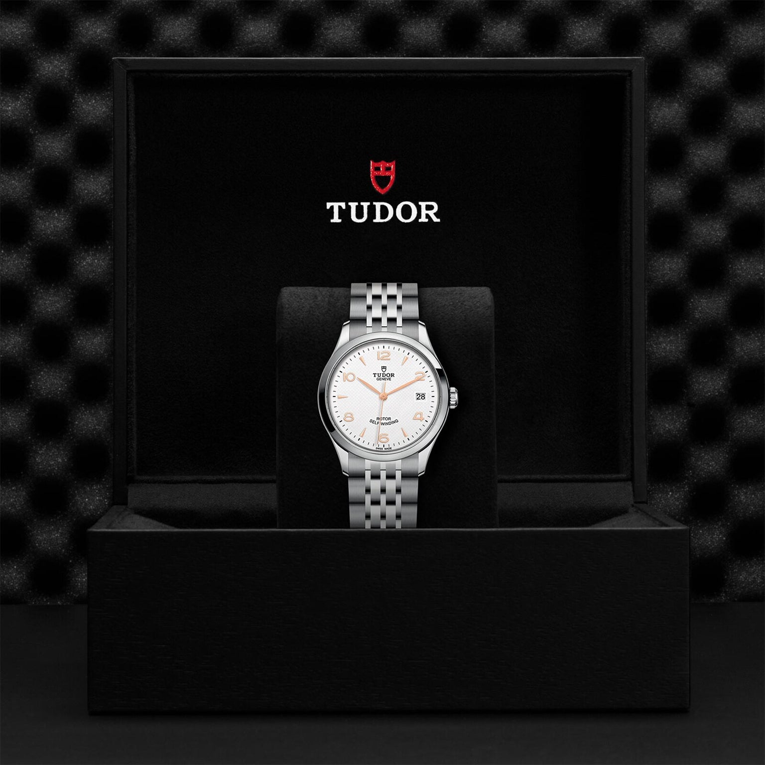 A TUDOR 1926 wristwatch with a white dial and silver metal band is showcased standing upright in a black TUDOR-branded presentation box, highlighting its classic elegance.