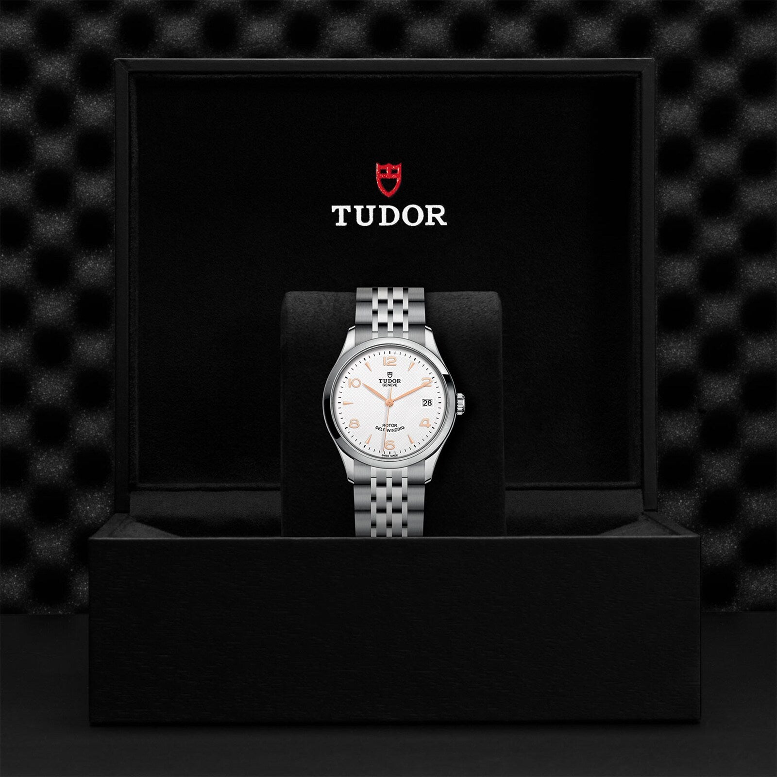 A TUDOR 1926 wristwatch with a white dial and silver metal band is showcased standing upright in a black TUDOR-branded presentation box, highlighting its classic elegance.