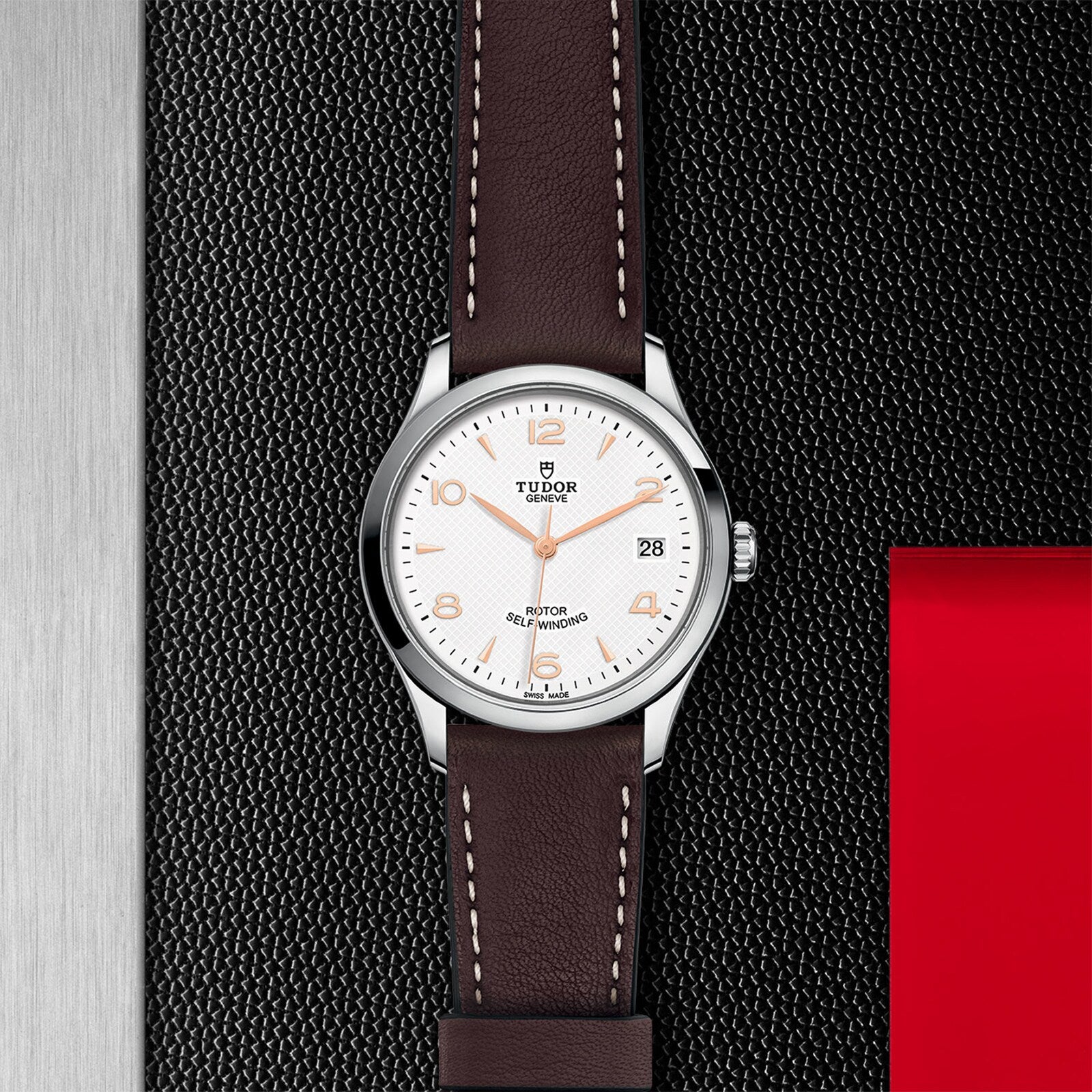 A TUDOR 1926 wristwatch featuring a white dial, brown leather strap, silver bezel, and date display rests on a textured black surface with red and metallic details—perfect for fans of classic elegance.