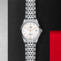 A TUDOR 1926 silver wristwatch with a white dial, orange Arabic numerals, and date window is displayed on a textured black and metallic background with a red accent, celebrating the timeless appeal of mechanical watches.