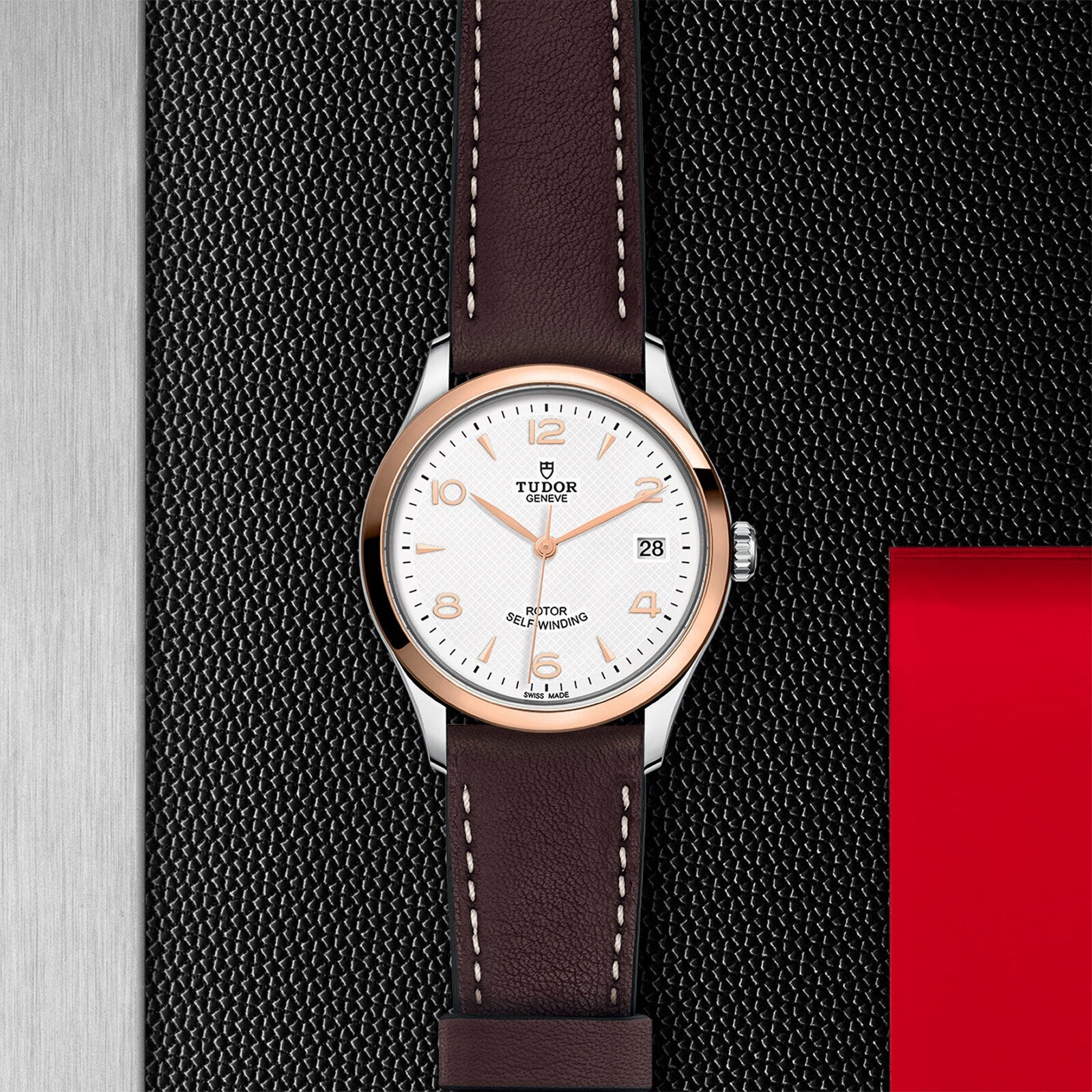 The TUDOR 1926 wristwatch features a white dial, brown leather strap, and silver case, elegantly displayed on a textured black surface with metallic and red accents.