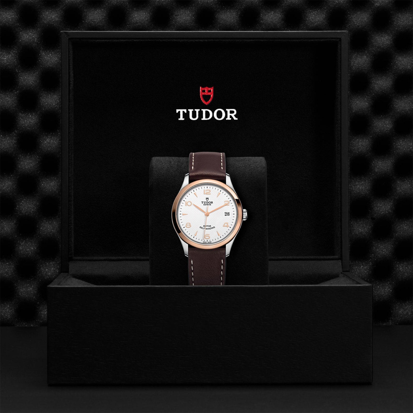 A TUDOR 1926 wristwatch with a white dial and brown leather strap rests in an open black TUDOR-branded box, showcasing the brand’s heritage of mechanical watchmaking.