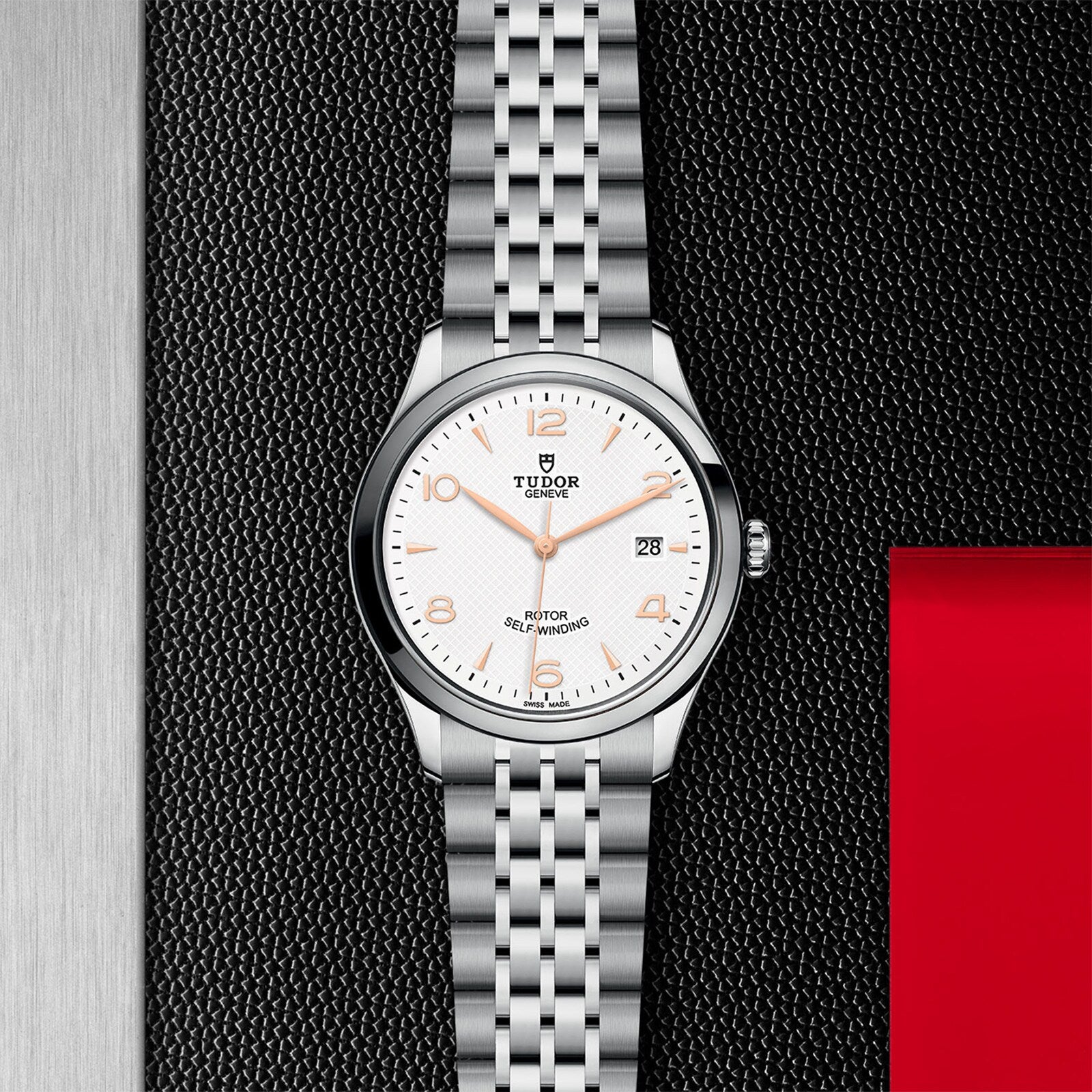 The TUDOR 1926 is a timeless silver wristwatch featuring a white dial, gold hour markers, date display, and metal bracelet, shown on a black and steel surface with a red accent. Perfect for lovers of mechanical watches.