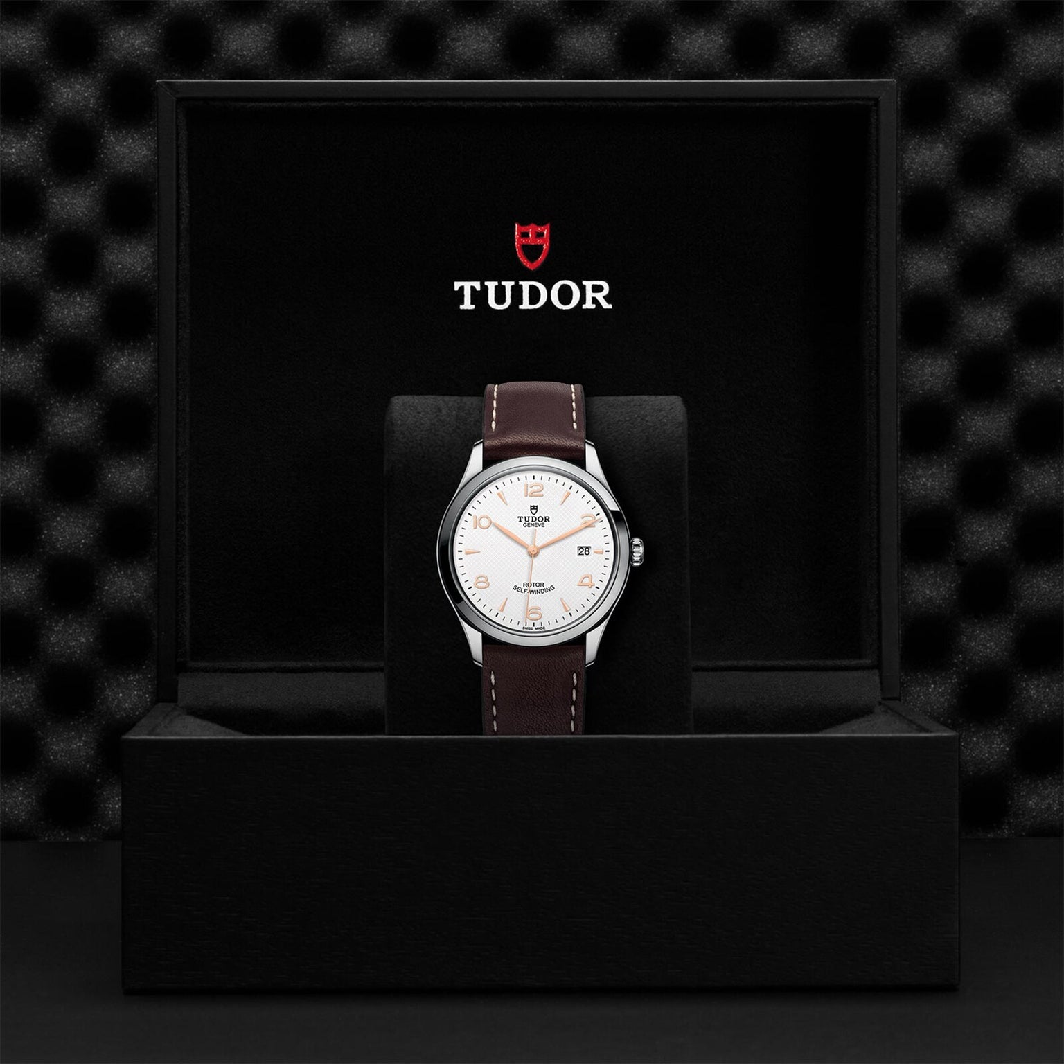 The TUDOR 1926 wristwatch with a white dial and brown leather strap is displayed upright in a black presentation box with the TUDOR logo on the inside lid, highlighting its classic and timeless mechanical design.