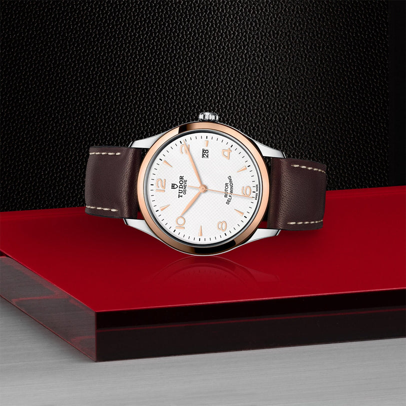 A TUDOR 1926 watch with a white dial, brown leather strap, and silver case is showcased on a red and black geometric surface against a textured black background, capturing the timeless elegance of mechanical timepieces.