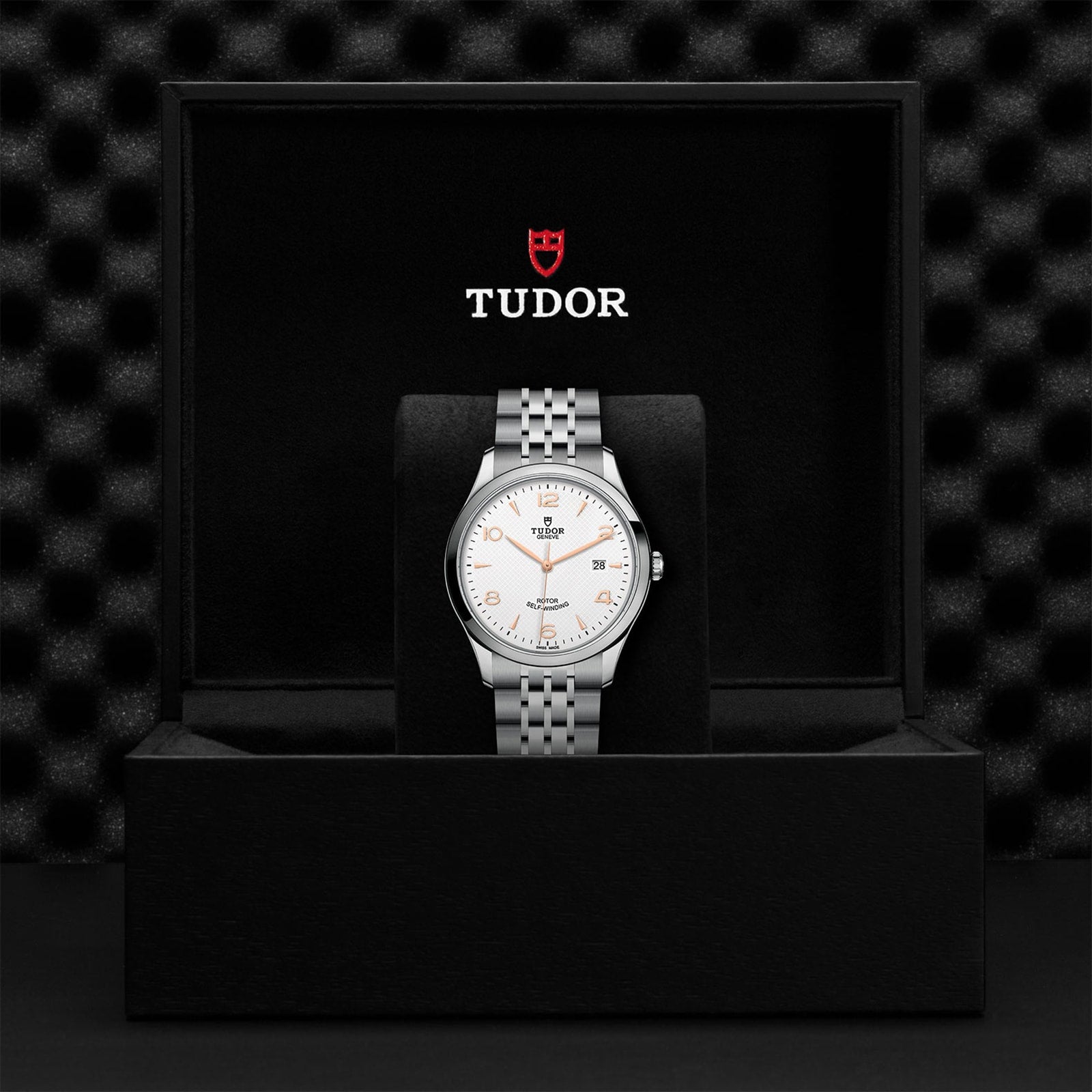 A silver TUDOR 1926 wristwatch with a white dial is showcased upright in an open black TUDOR box against a textured black background, reflecting the mechanical watch heritage linked to Rolex’s founder.