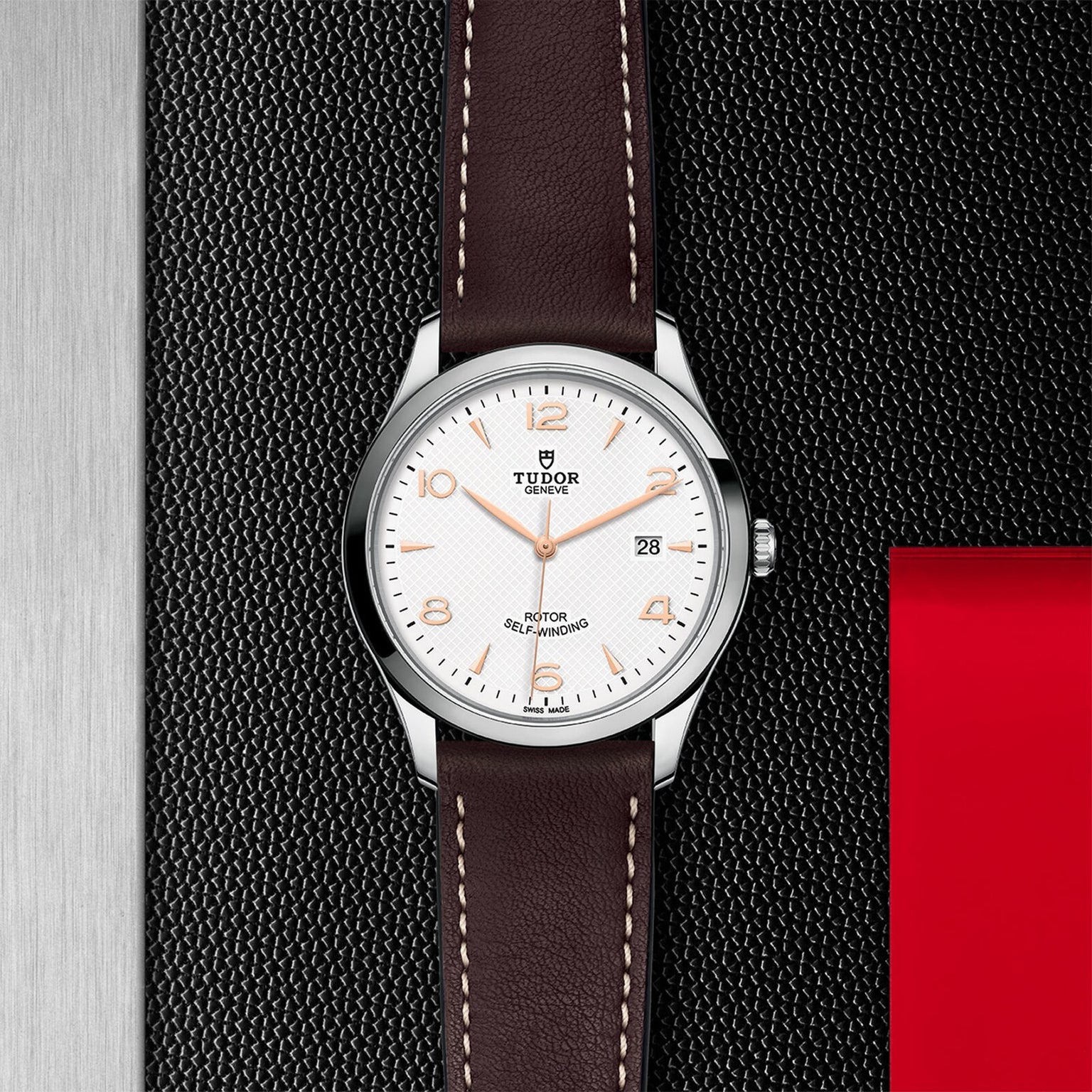 A TUDOR 1926 wristwatch featuring a white dial, brown leather strap, and silver case is displayed on a textured black and metallic background with a red accent, highlighting the refined style of TUDOR.