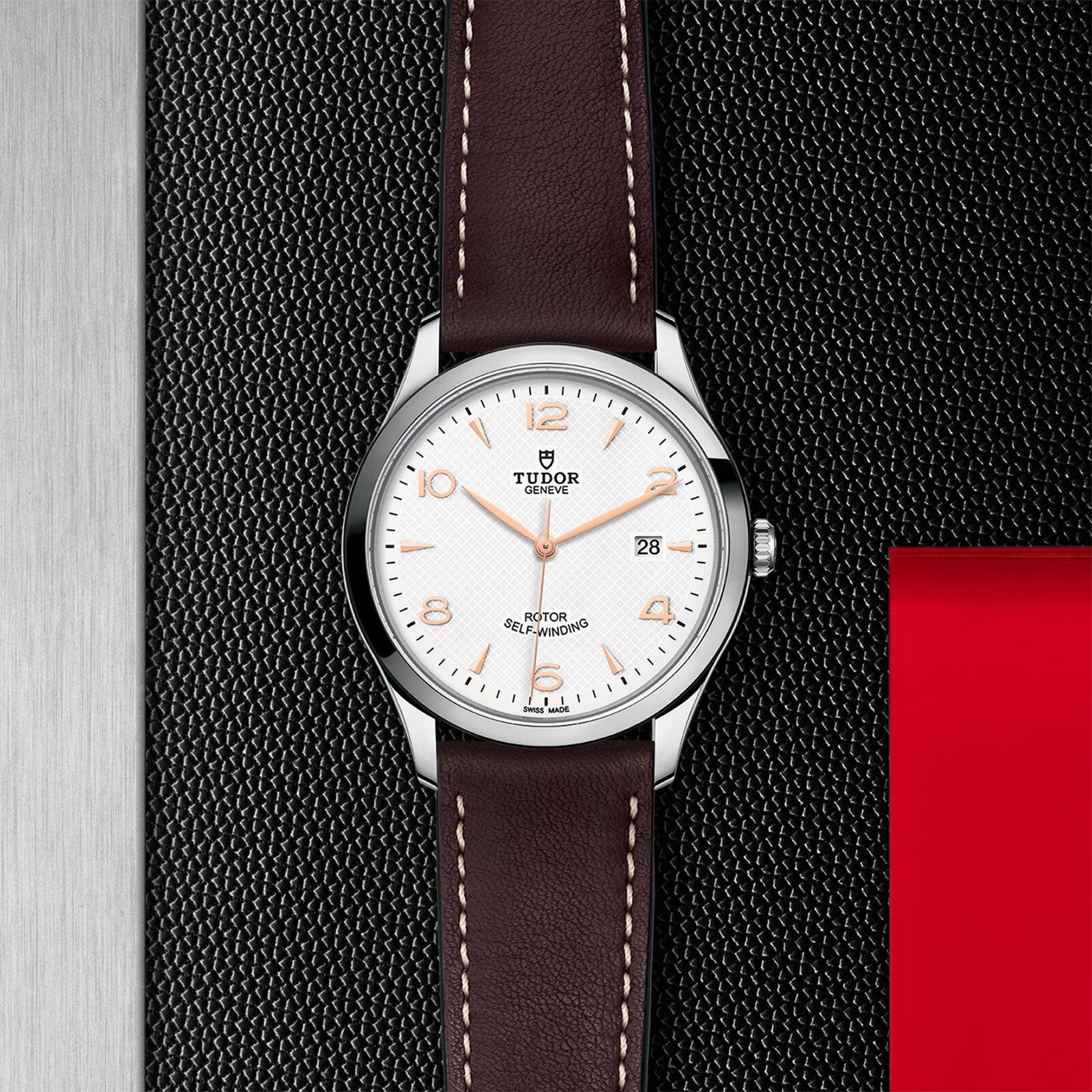 A TUDOR 1926 wristwatch featuring a white dial, brown leather strap, and silver case is displayed on a textured black and metallic background with a red accent, highlighting the refined style of TUDOR.