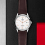 A TUDOR 1926 wristwatch featuring a white dial, brown leather strap, and silver case is displayed on a textured black and metallic background with a red accent, highlighting the refined style of TUDOR.
