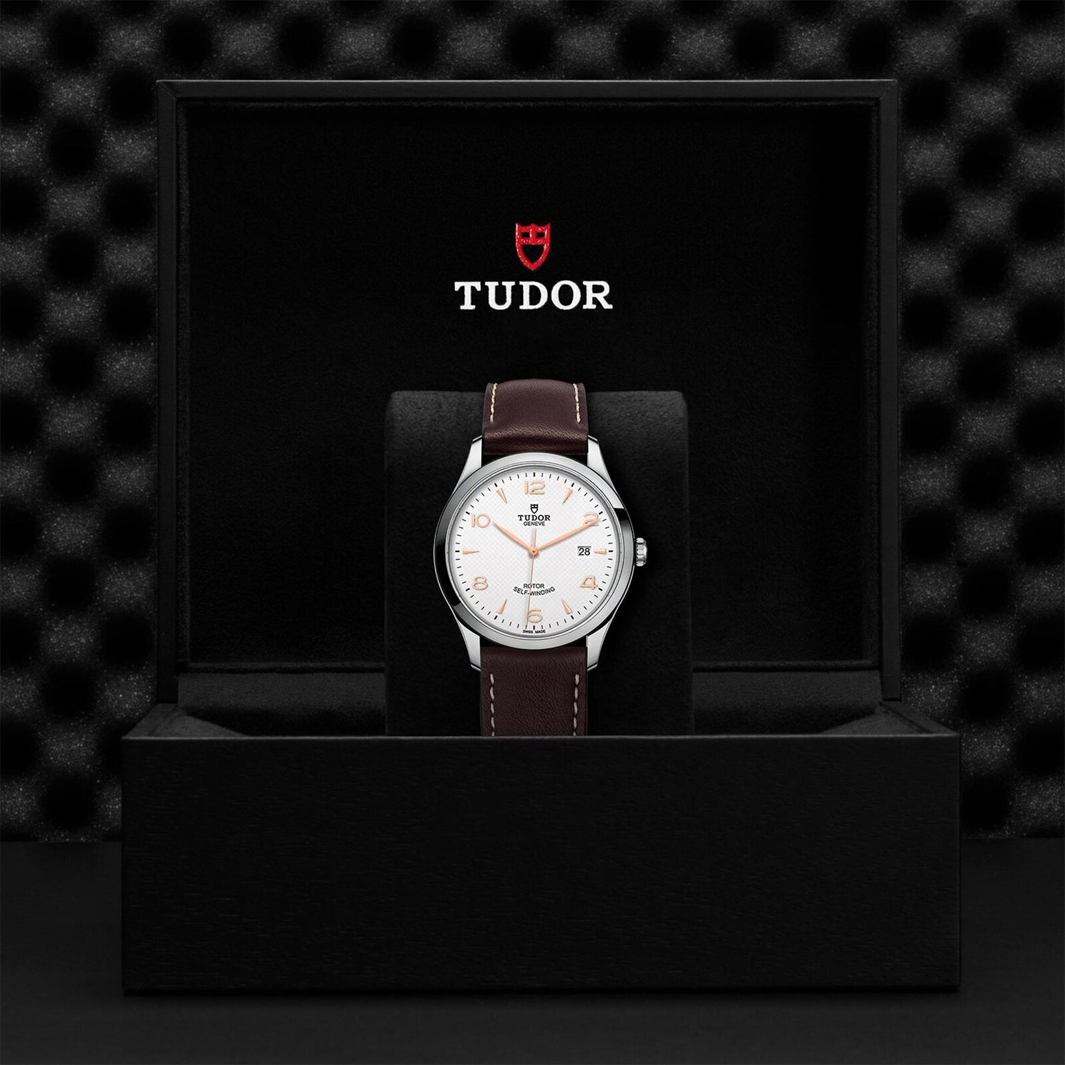 A TUDOR 1926 wristwatch with a brown leather strap is displayed upright in an open black box with the TUDOR logo above—showcasing the elegant design and renowned Swiss craftsmanship of TUDOR watches.