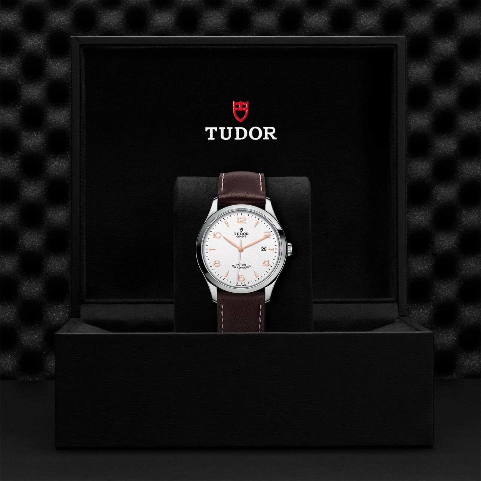 A TUDOR 1926 wristwatch with a brown leather strap is displayed upright in an open black box with the TUDOR logo above—showcasing the elegant design and renowned Swiss craftsmanship of TUDOR watches.