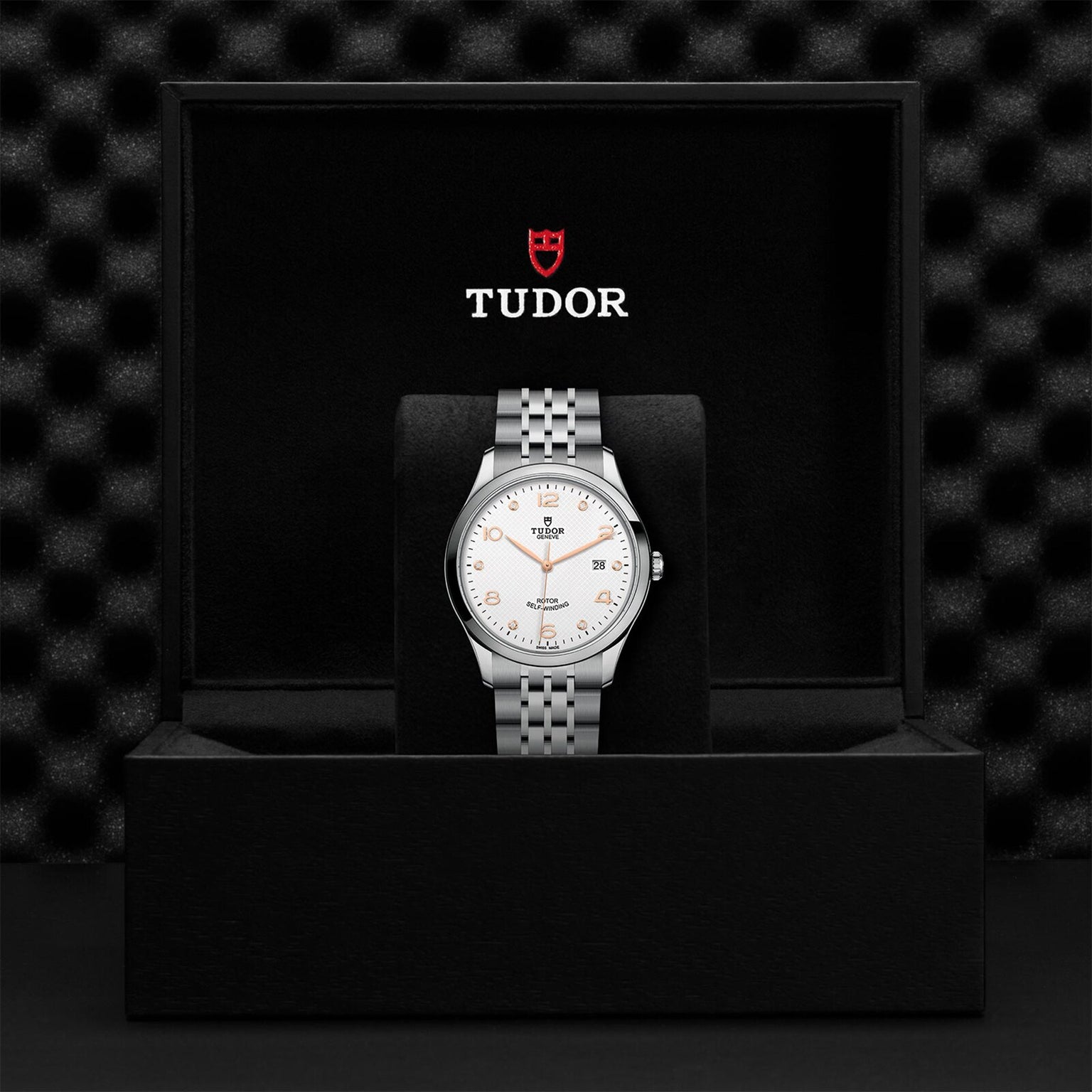 The TUDOR 1926 wristwatch, featuring a silver metal band and white dial, is displayed upright in a black presentation box with the TUDOR logo on the inside lid, reflecting the brand’s legacy in mechanical watchmaking.