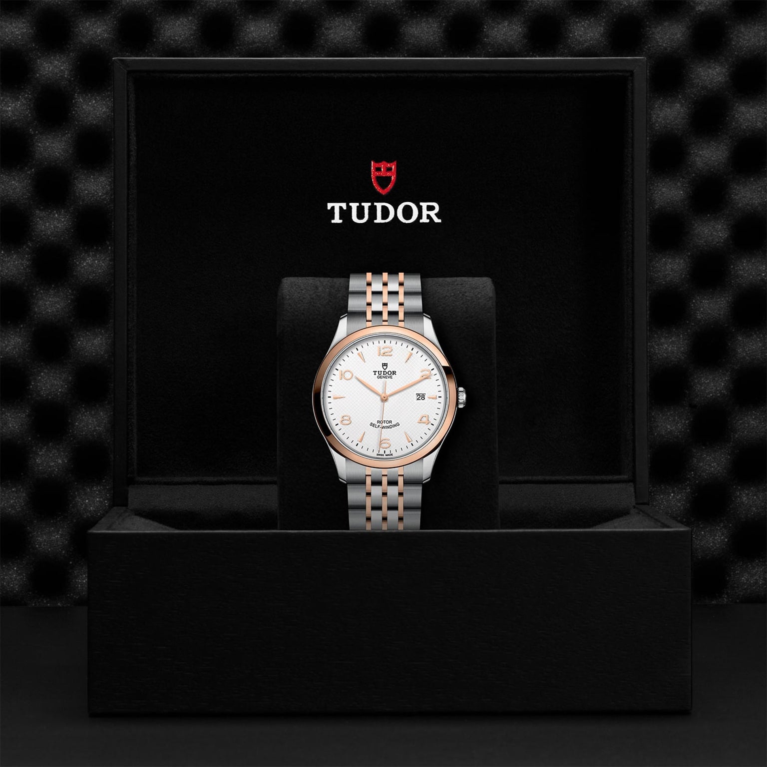 A TUDOR 1926 wristwatch with a silver and gold bracelet and white dial stands upright in a black TUDOR presentation box, emphasizing the brand’s heritage in mechanical timepieces.