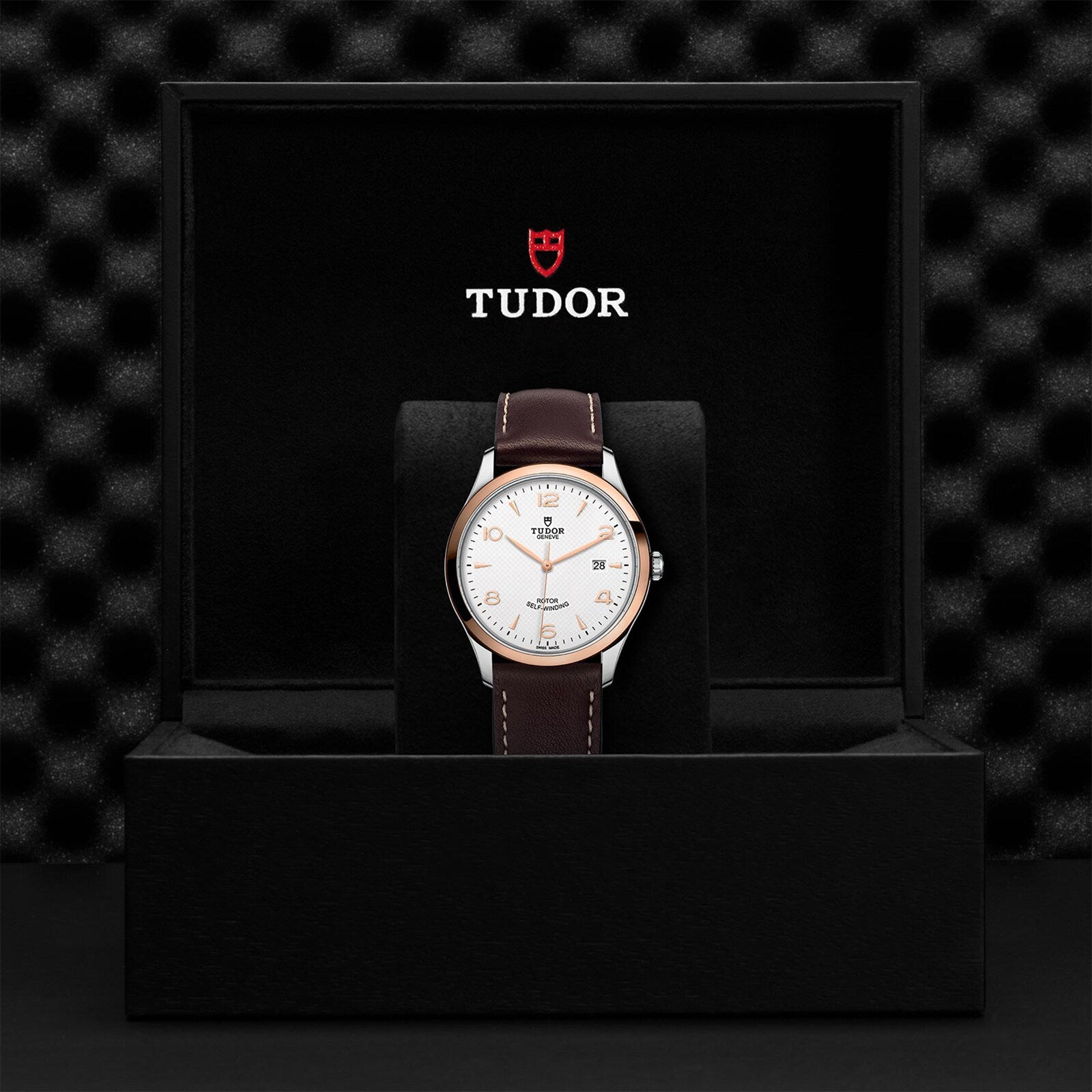 A TUDOR 1926 wristwatch with a brown leather strap and white dial stands upright in an open black TUDOR box, showcasing refined mechanical watch craftsmanship.