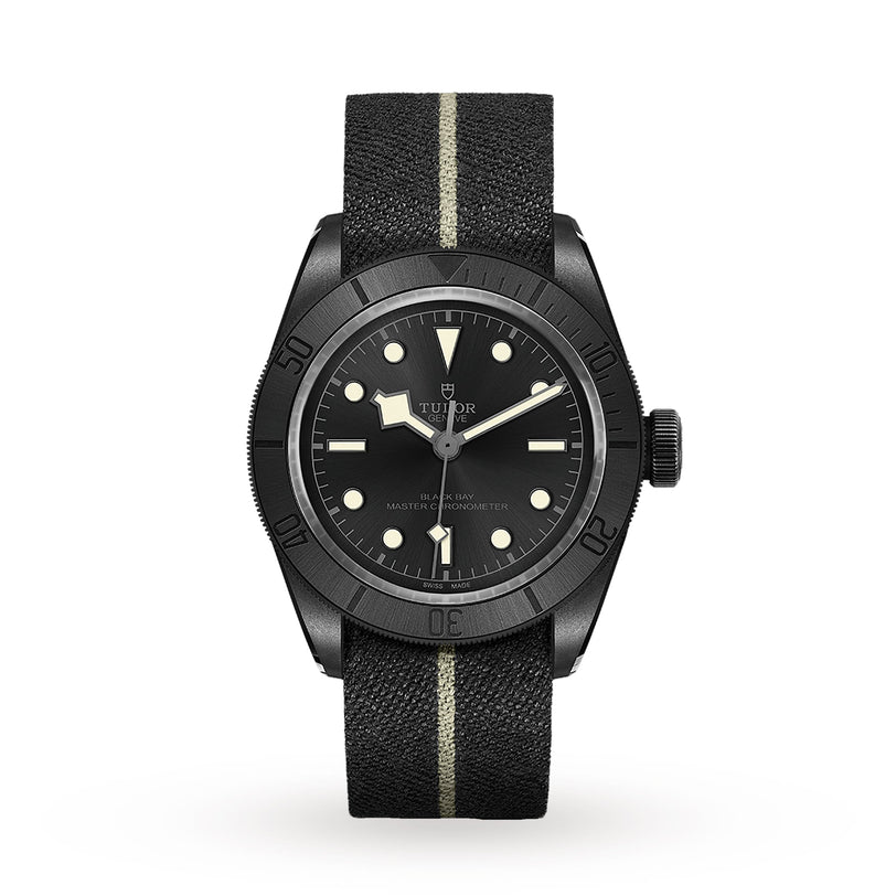 The TUDOR Black Bay Ceramic is a Swiss-made wristwatch featuring a black dial, luminous markers, black bezel, and a black fabric strap with a single beige stripe.