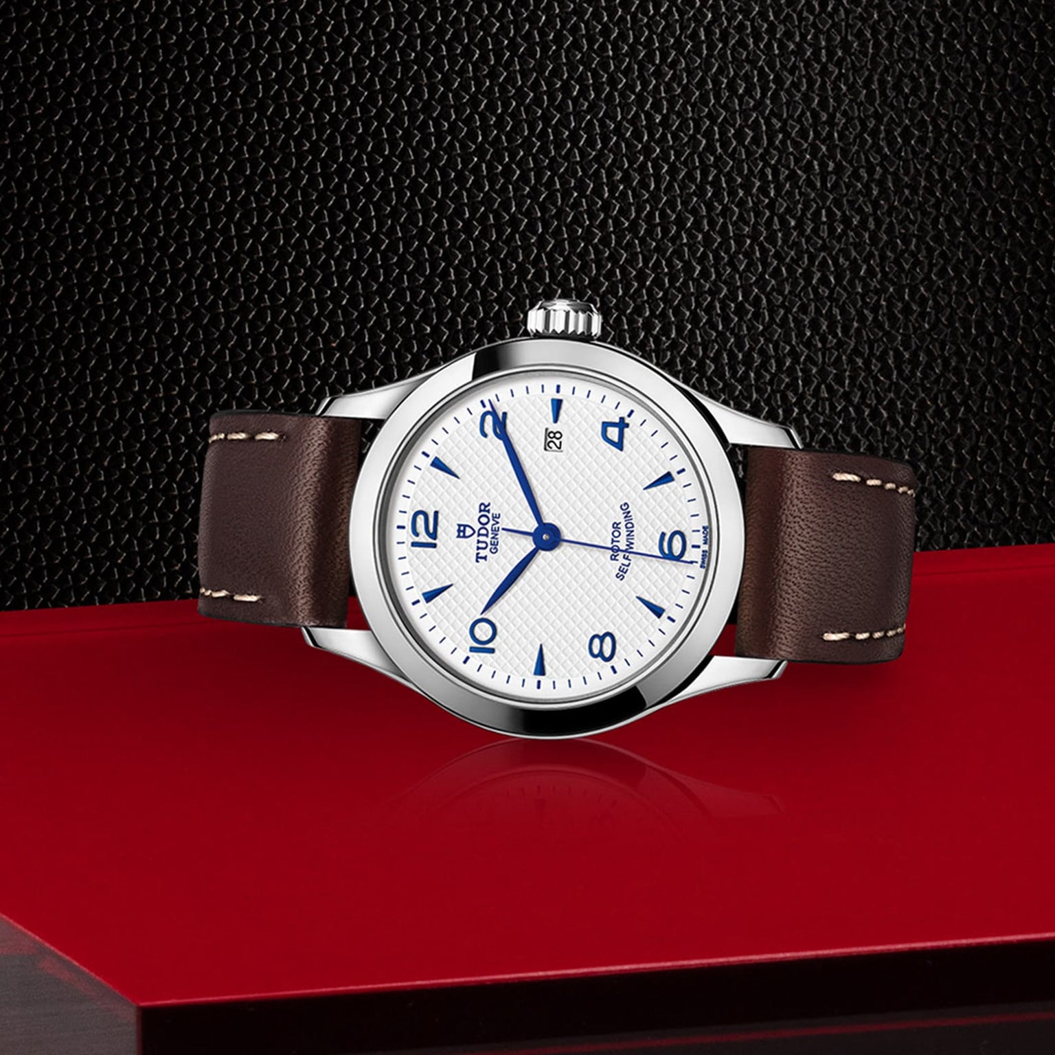 A TUDOR 1926 wristwatch with a white textured dial, blue numerals and hands, and a brown leather strap is displayed on a red surface against a black textured background.