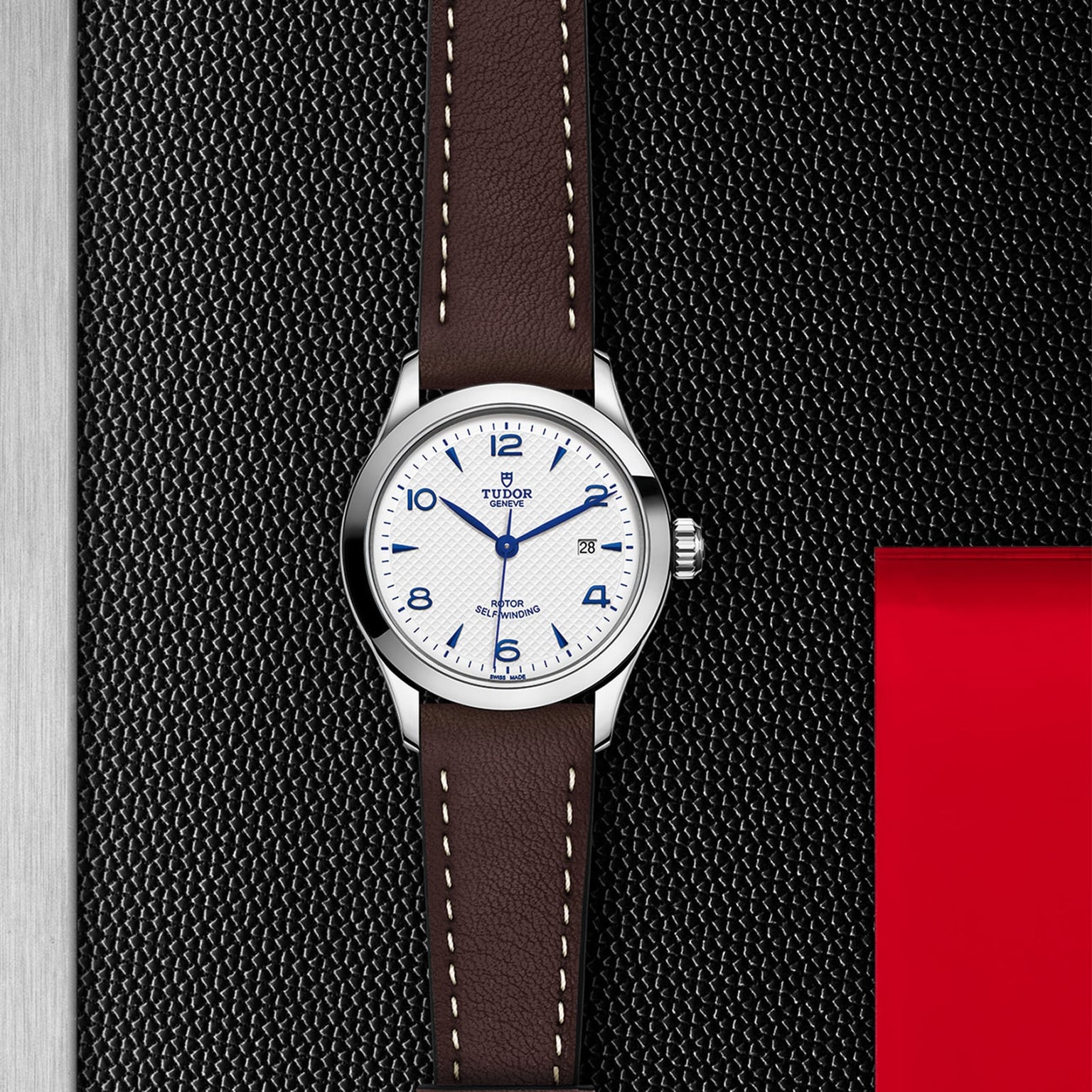 A TUDOR 1926 wristwatch with a white dial, blue numerals, brown leather strap, and date display rests on a textured black and red surface—a timeless mechanical watch from TUDOR.