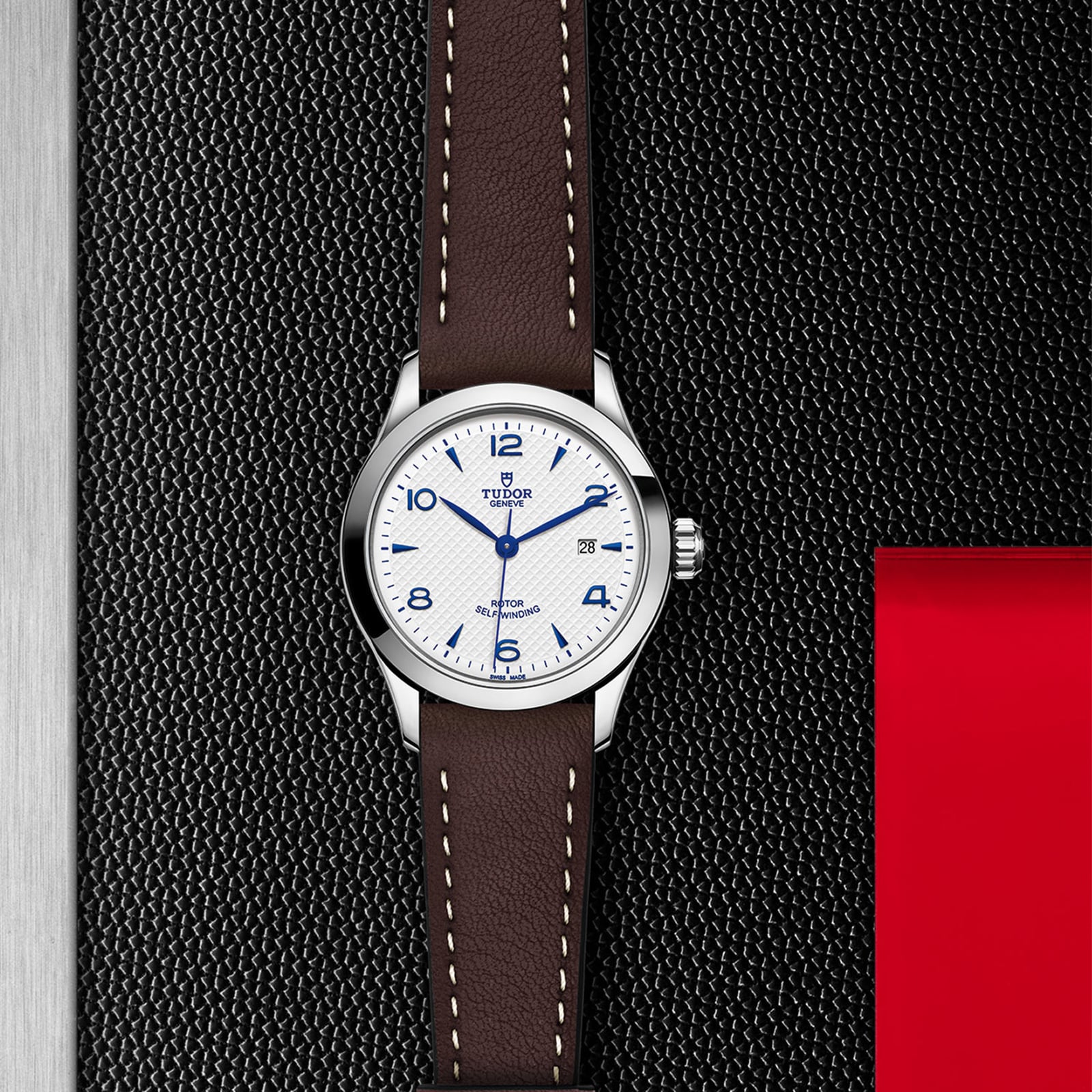 A TUDOR 1926 wristwatch with a white dial, blue numerals, brown leather strap, and date display rests on a textured black and red surface—a timeless mechanical watch from TUDOR.