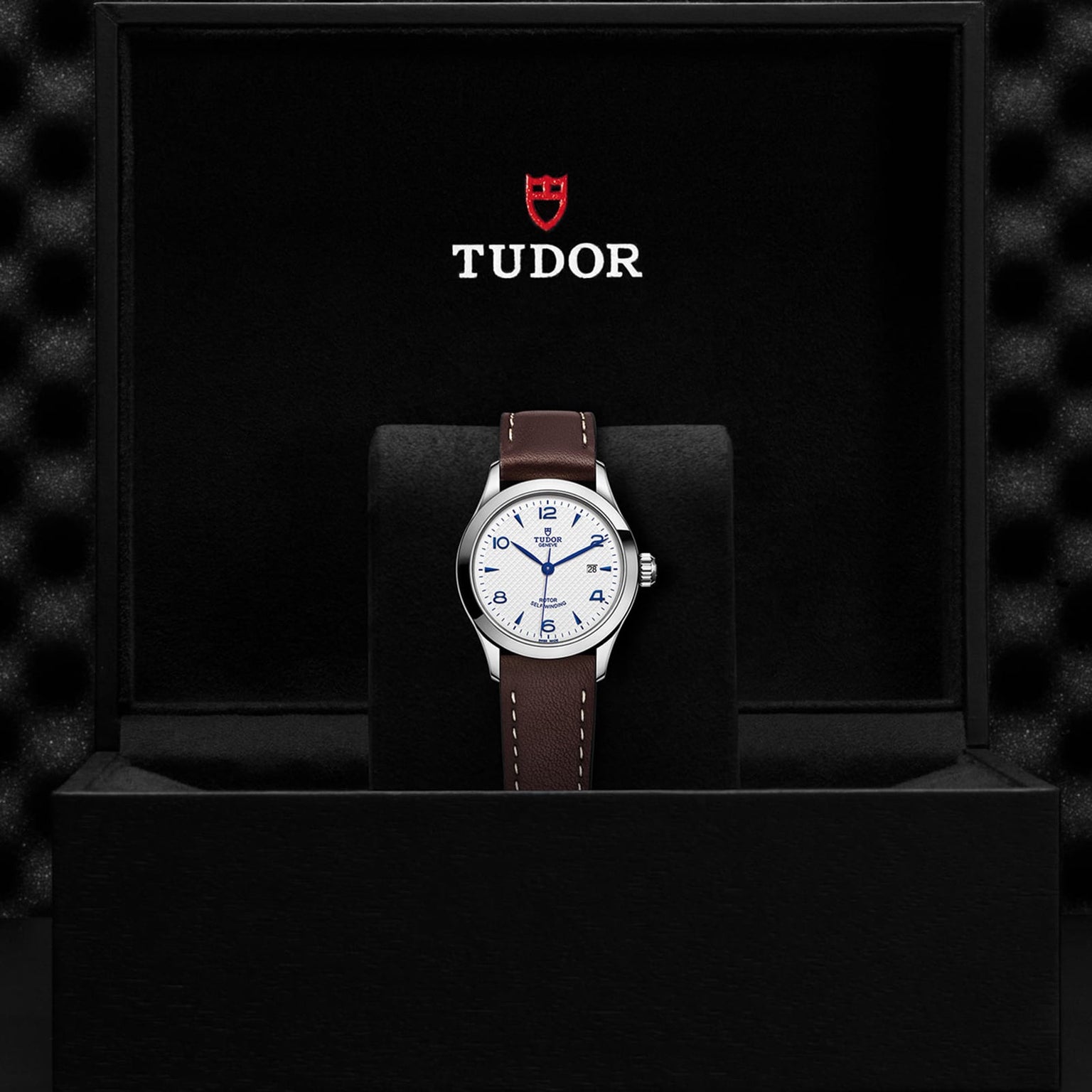 A TUDOR 1926 wristwatch with a white dial and brown leather strap is displayed in a black box with the TUDOR logo and shield, highlighting the refined style of this mechanical timepiece.