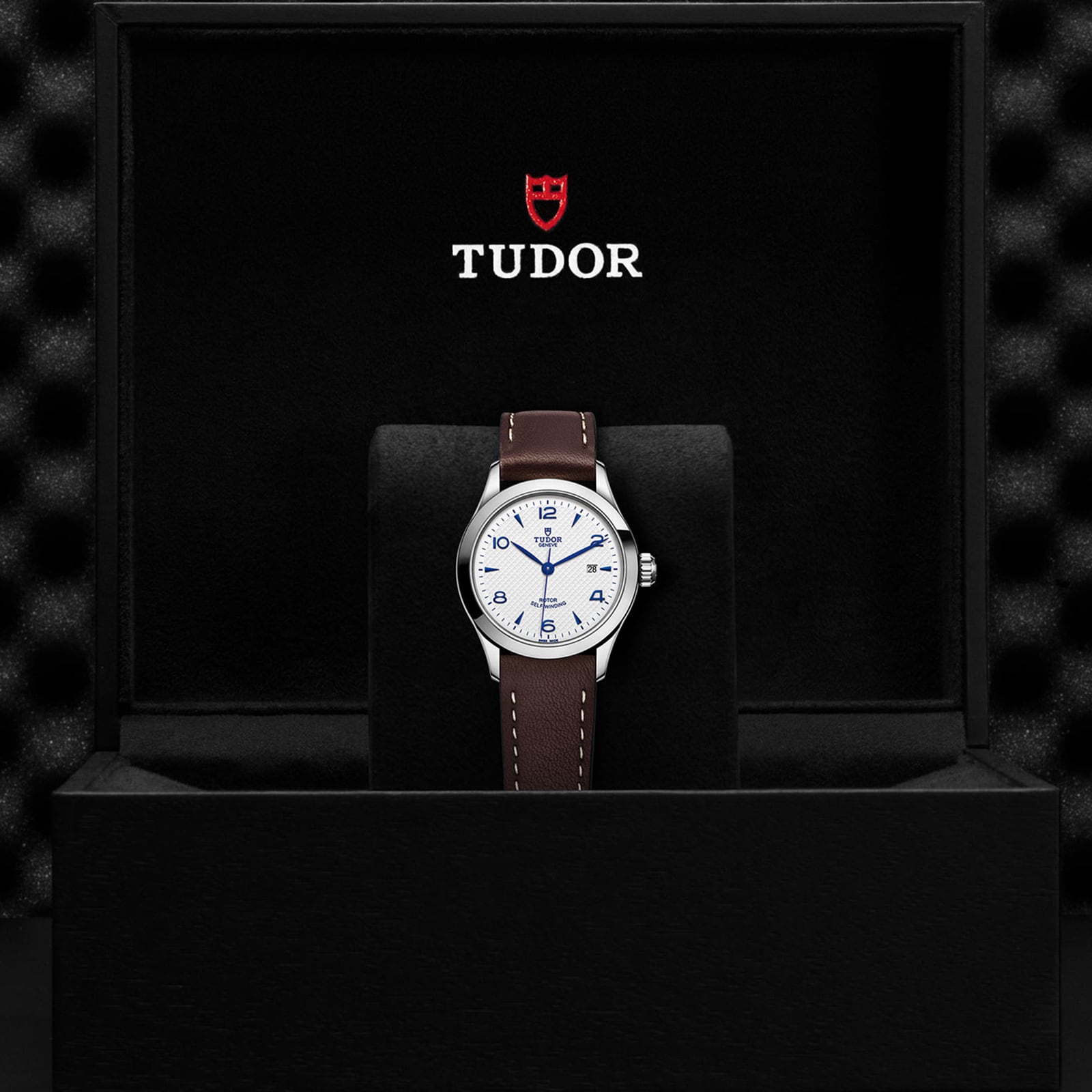A TUDOR 1926 wristwatch with a white dial and brown leather strap is displayed in a black box with the TUDOR logo and shield, highlighting the refined style of this mechanical timepiece.