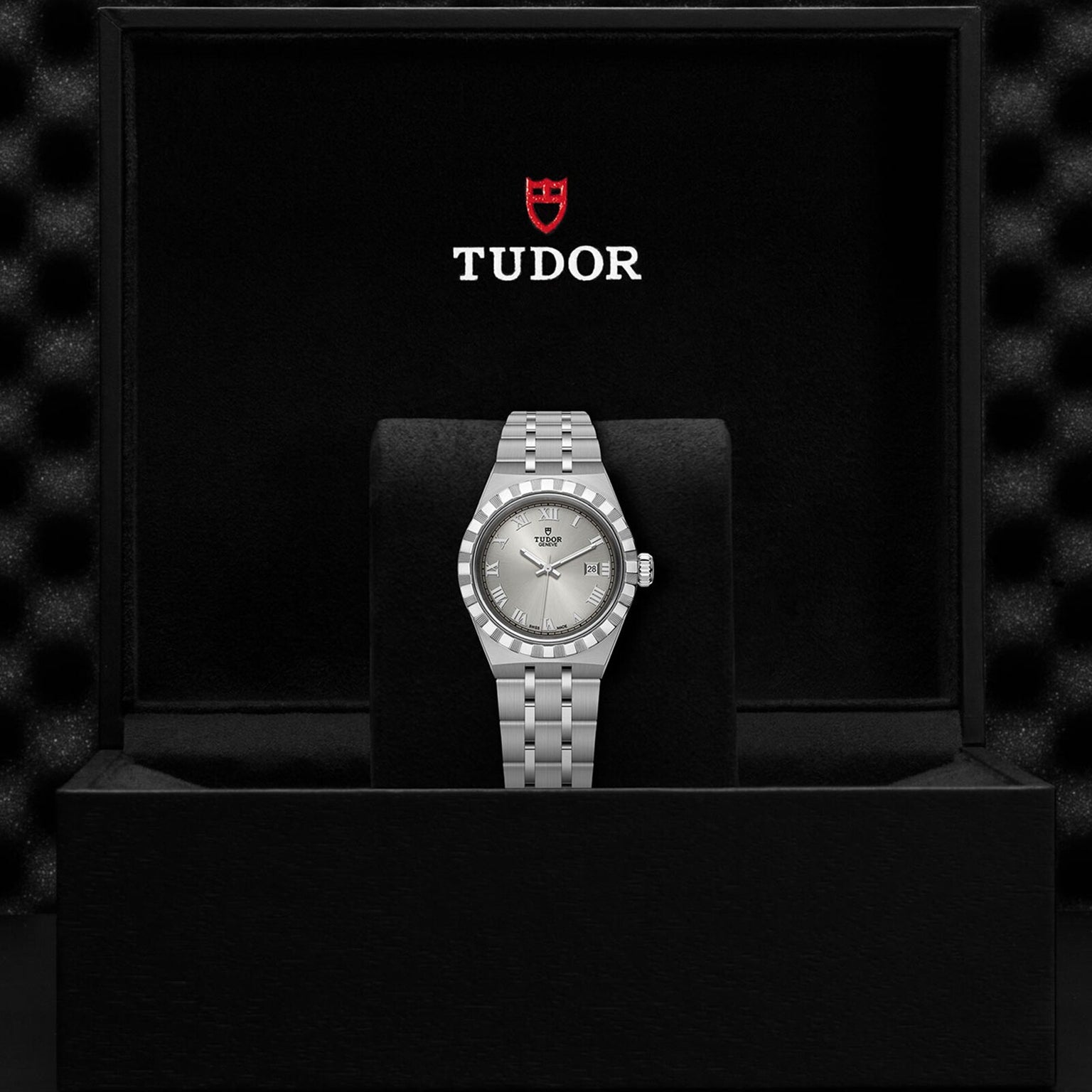 A silver TUDOR Royal wristwatch with a metal bracelet stands upright in a black presentation box, showcasing the TUDOR logo and name inside the lid—an elegant example of sport-chic timepieces.