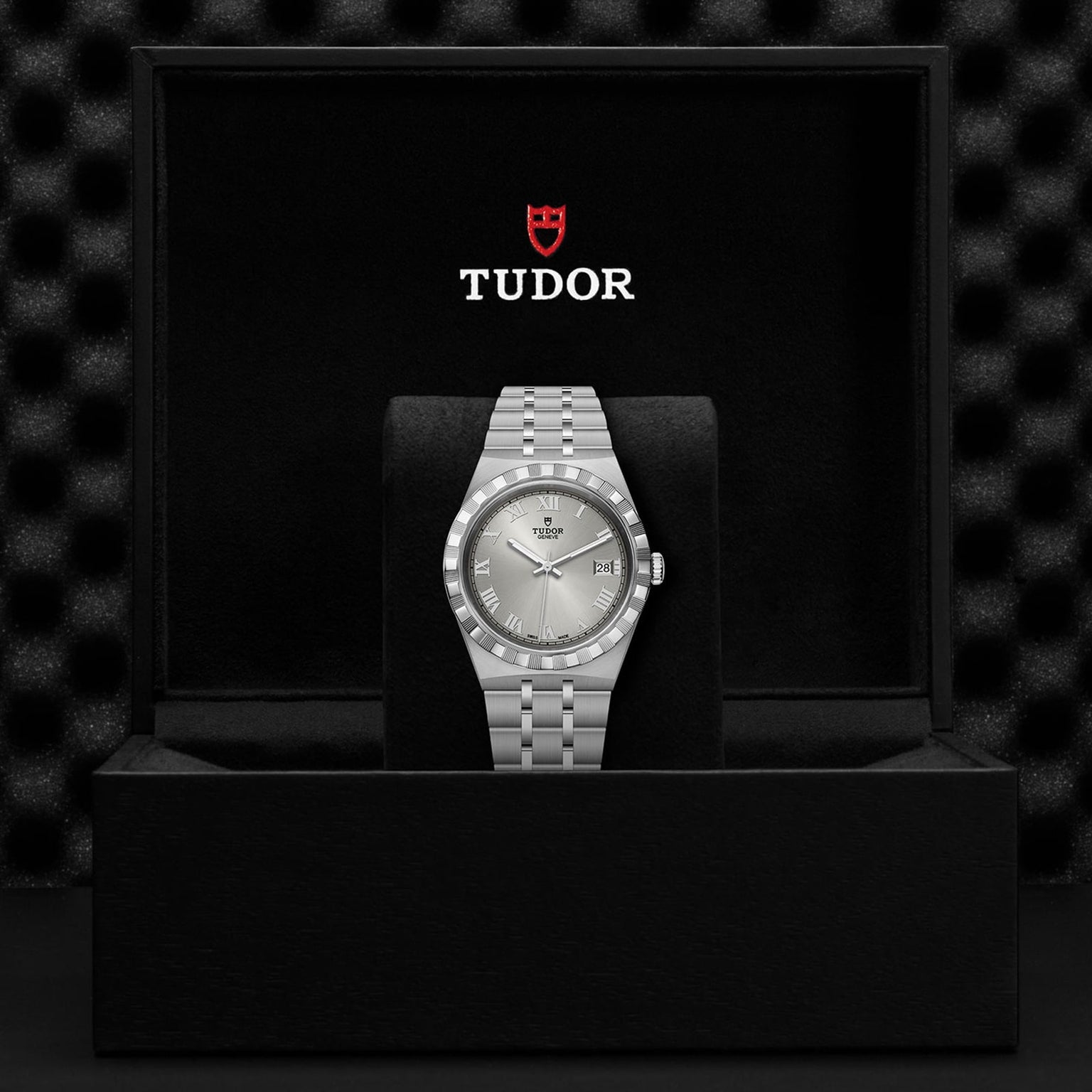 A TUDOR Royal wristwatch is upright in a black presentation box featuring the Tudor logo.