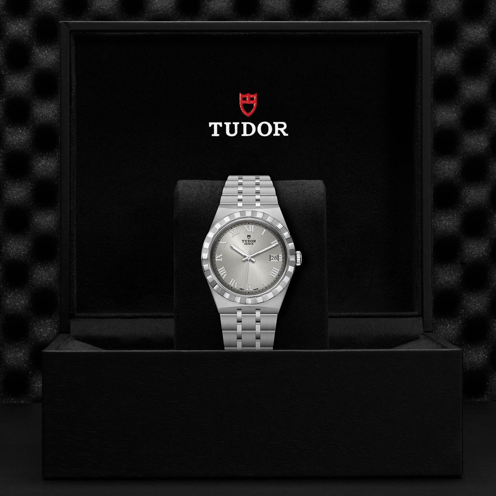 A TUDOR Royal wristwatch is upright in a black presentation box featuring the Tudor logo.