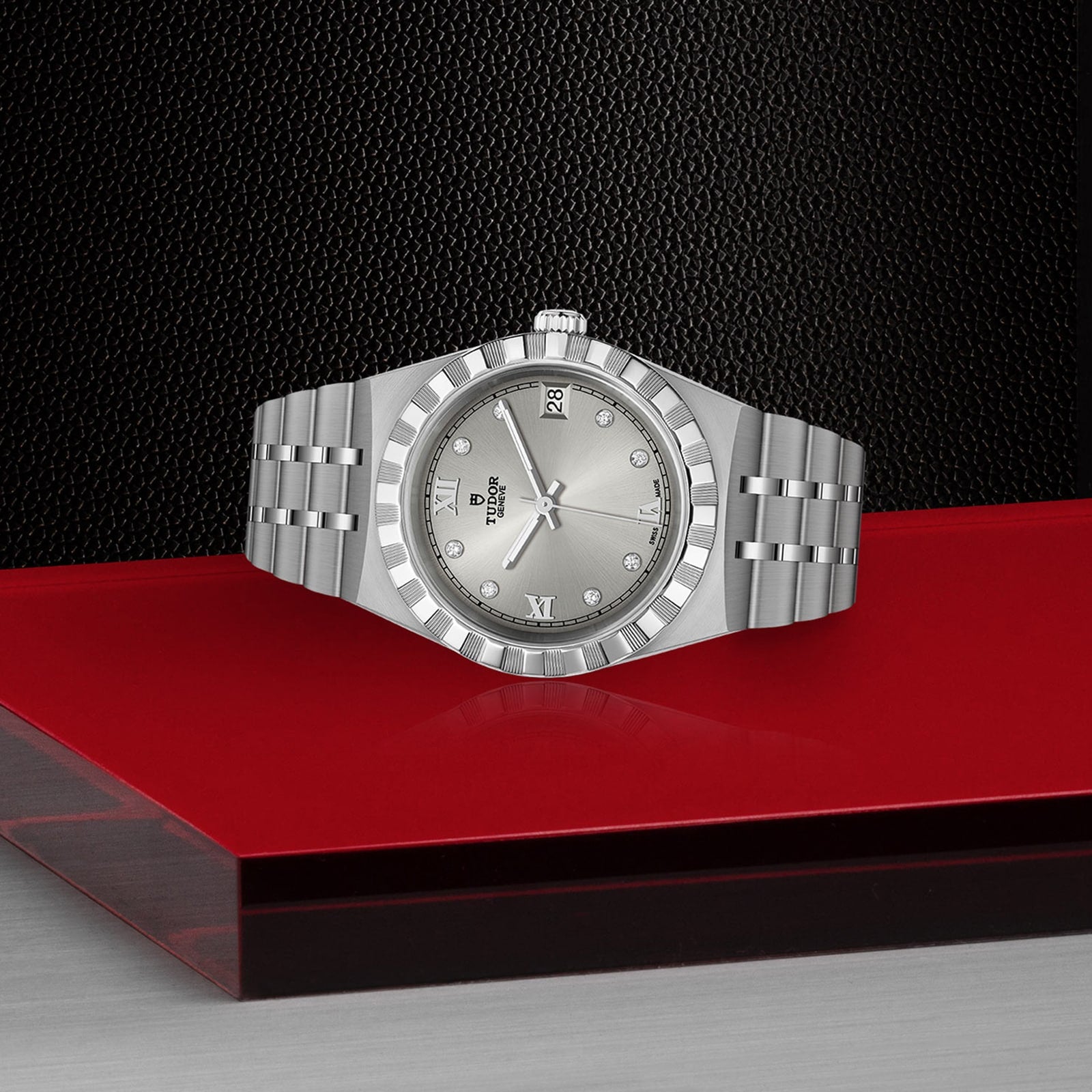 A TUDOR Royal wristwatch with a silver metallic band and diamond hour markers is displayed on a red and gray platform against a textured black background, exemplifying the sport-chic style of automatic watches.