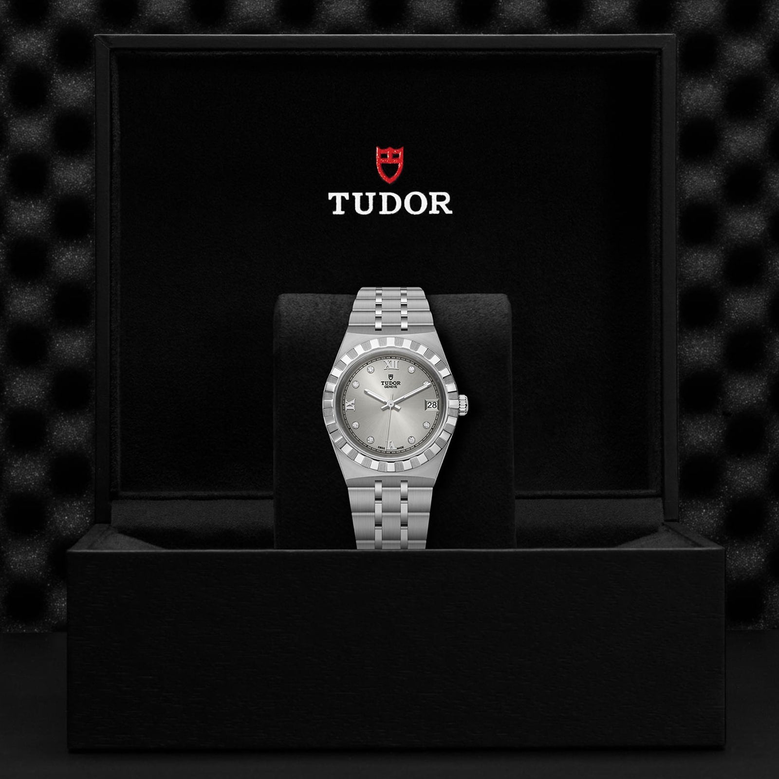 A TUDOR Royal wristwatch with a silver-toned metal bracelet is showcased upright in an open black TUDOR watch box against a dark background, capturing the refined, sport-chic style of this automatic timepiece.