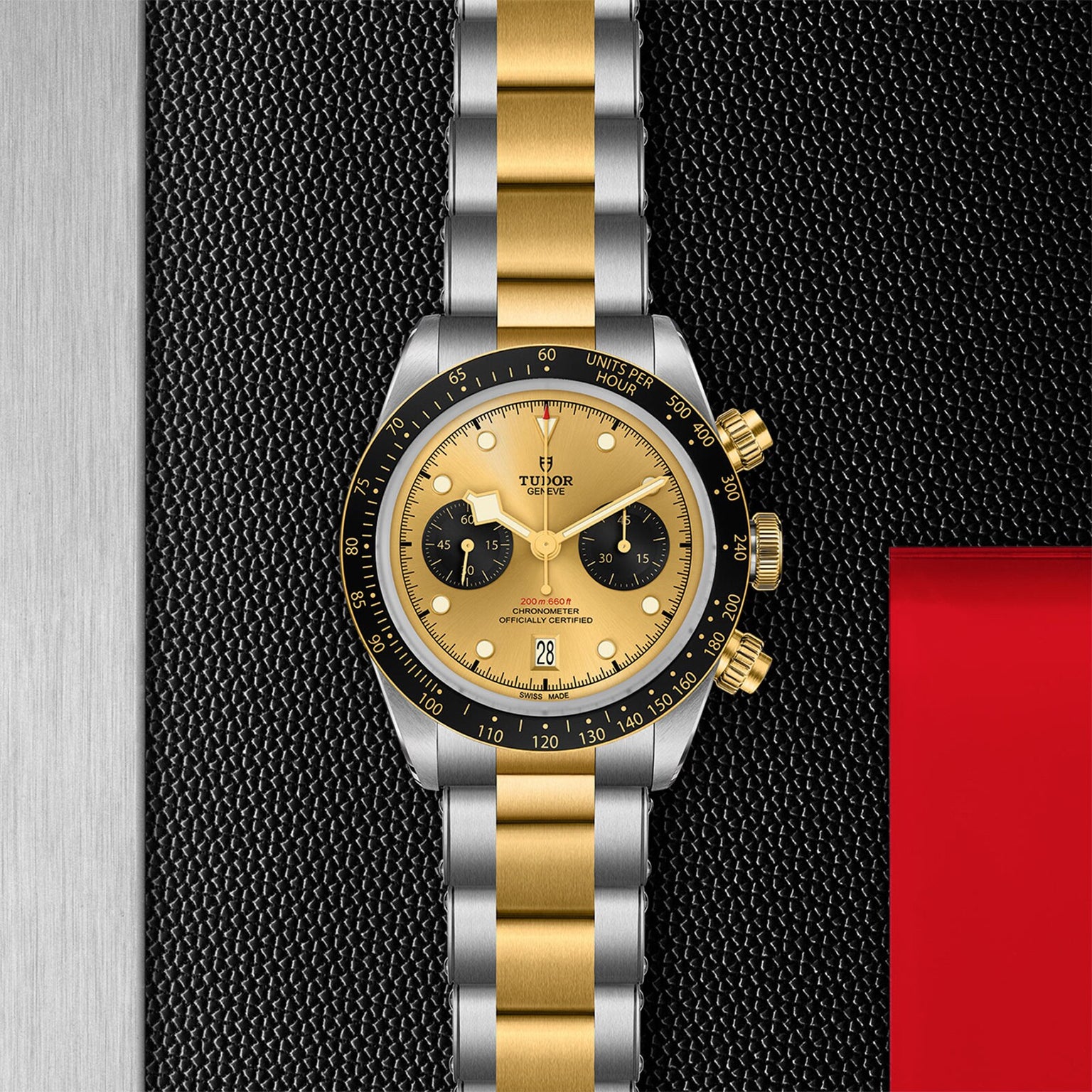 The TUDOR Black Bay Chrono S&G wristwatch features a two-tone yellow gold and silver bracelet, black bezel, gold dial with two black subdials, and is displayed on a textured black surface.