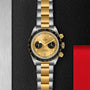 The TUDOR Black Bay Chrono S&G wristwatch features a two-tone yellow gold and silver bracelet, black bezel, gold dial with two black subdials, and is displayed on a textured black surface.