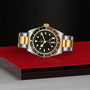 The TUDOR Black Bay GMT S&G wristwatch, featuring yellow gold accents and a black dial and bezel, is showcased on a red and gray platform against a textured black background.