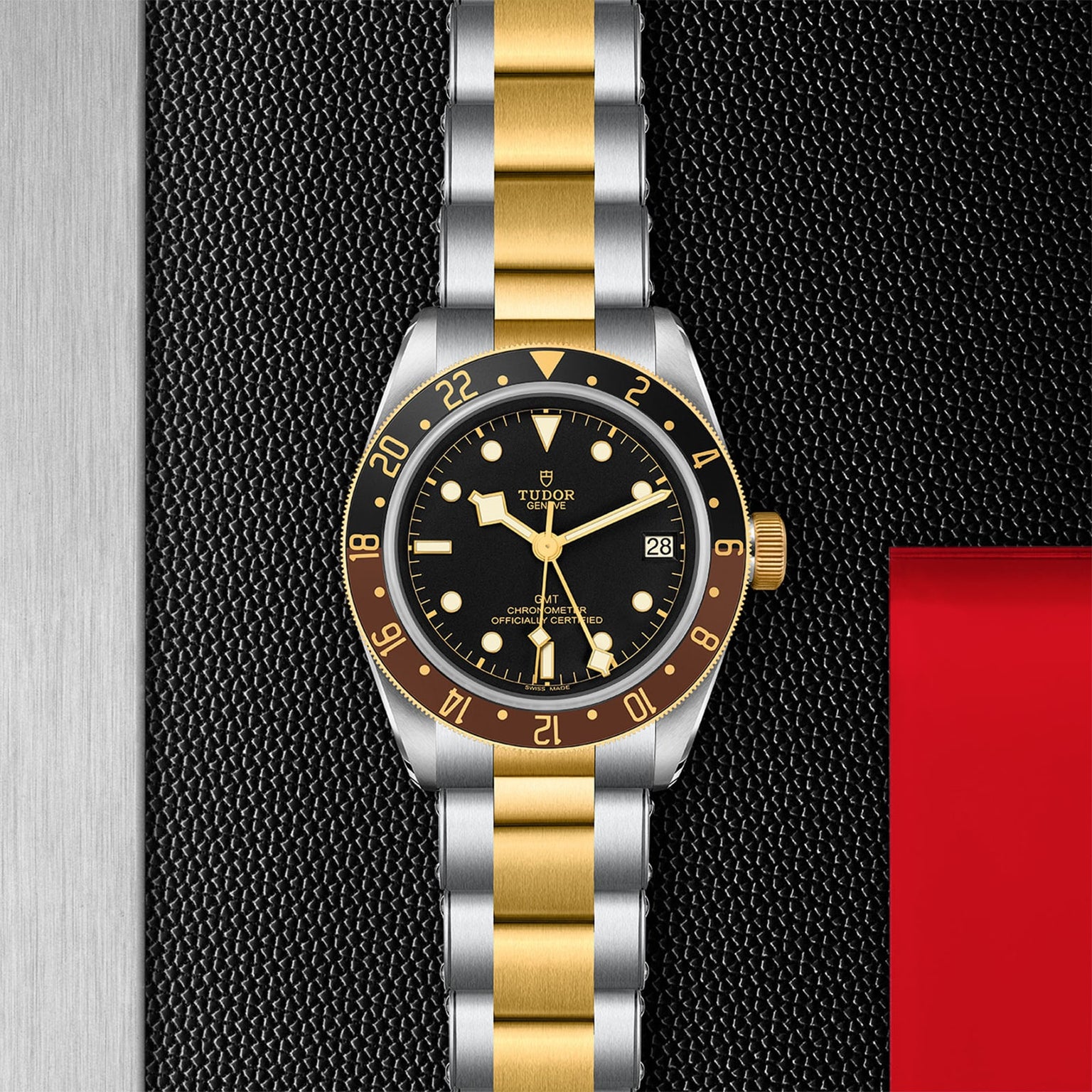 The TUDOR Black Bay GMT S&G wristwatch, featuring a black dial, yellow gold bezel, and sleek silver-and-gold bracelet, is displayed on a textured black surface.