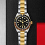 The TUDOR Black Bay GMT S&G wristwatch, featuring a black dial, yellow gold bezel, and sleek silver-and-gold bracelet, is displayed on a textured black surface.