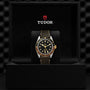 A TUDOR Black Bay GMT S&G wristwatch with a black and yellow gold bezel is displayed in an open black presentation box featuring the TUDOR logo, highlighting the heritage of TUDOR divers’ watches.
