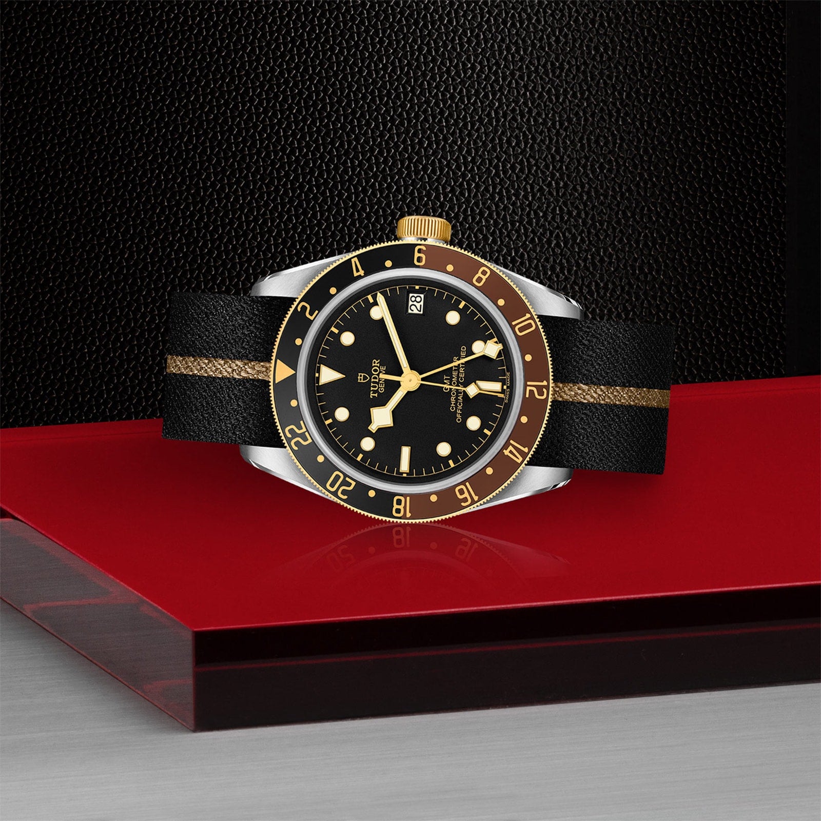 The TUDOR Black Bay GMT S&G luxury watch, inspired by Tudor divers’ models, showcases a black and gold face and a black strap with a gold stripe, displayed on red and gray surfaces against a textured black background.